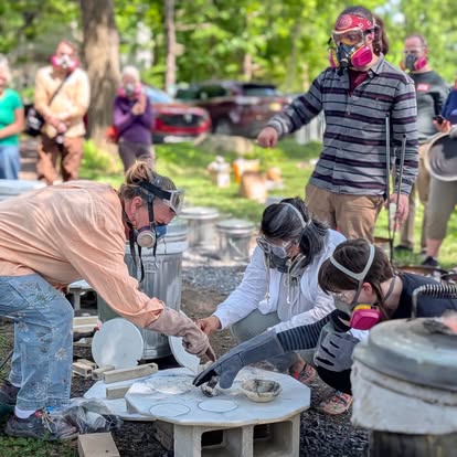 People wearing masks gather outdoors around a table with food, with trees and parked cars in the background.