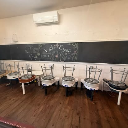 Chairs stacked upside down along a wall with a chalkboard and air conditioner above, wooden floor below.