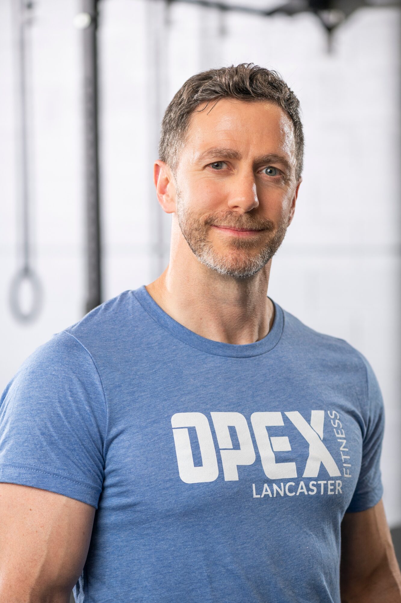Man with short dark hair and beard wearing a blue T-shirt with white text, standing indoors with gym equipment in background.