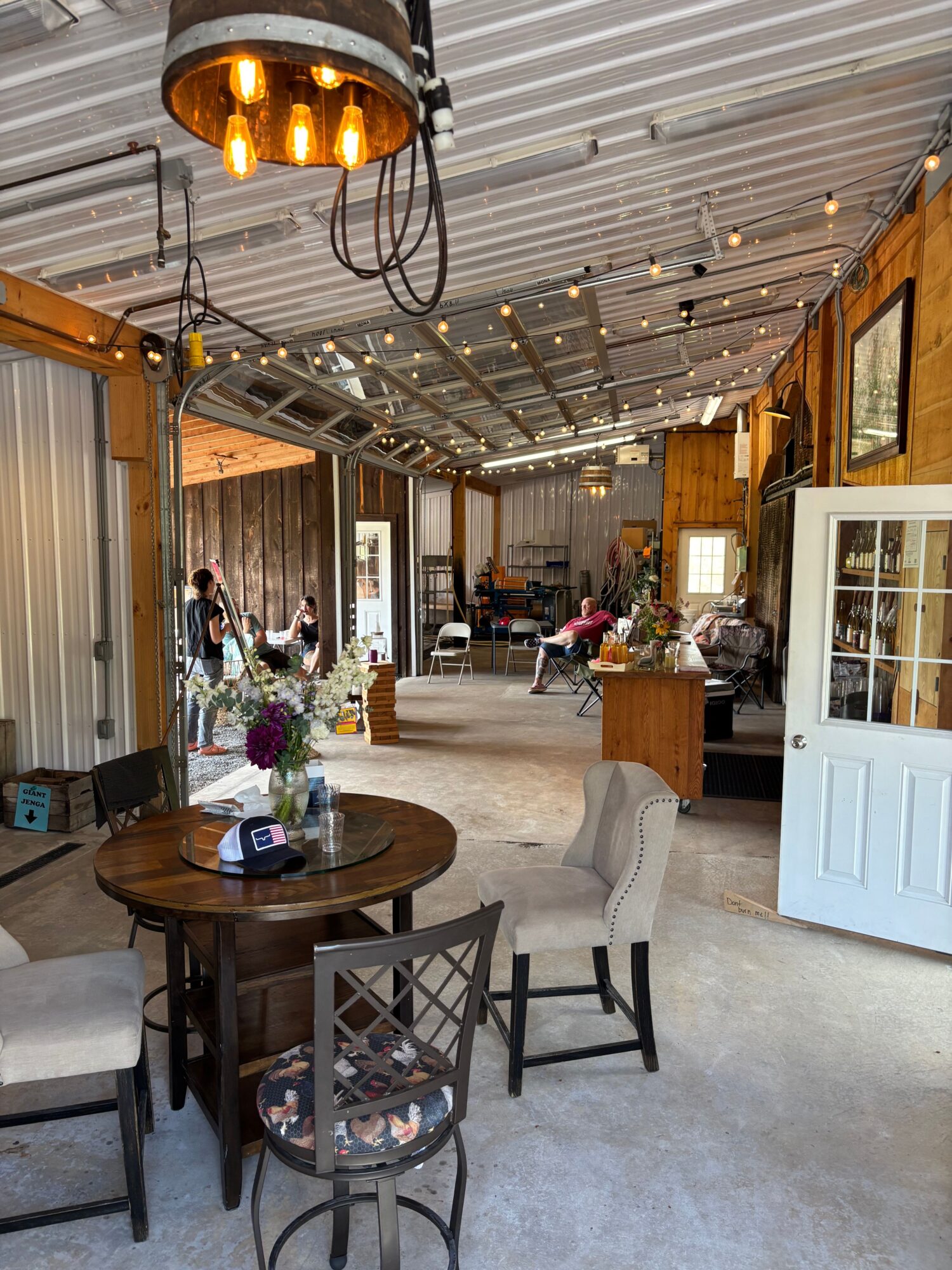 Indoor space with a round table, chairs, and a display cabinet, decorated with flowers and string lights, viewed from the side.