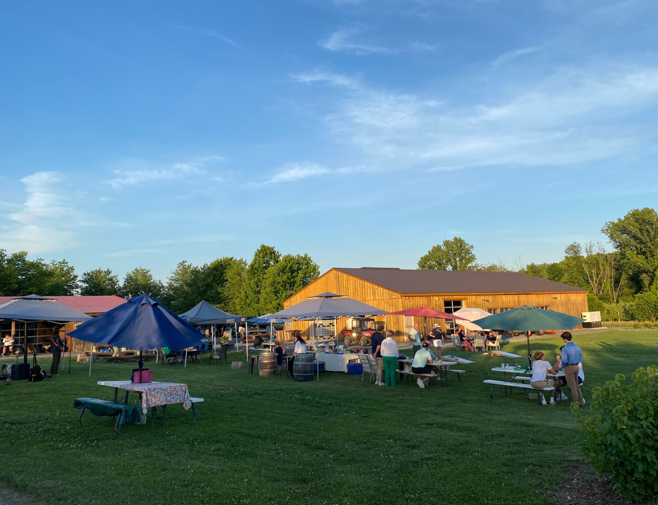 Outdoor gathering with tents, tables, and people on a grassy field under a blue sky.