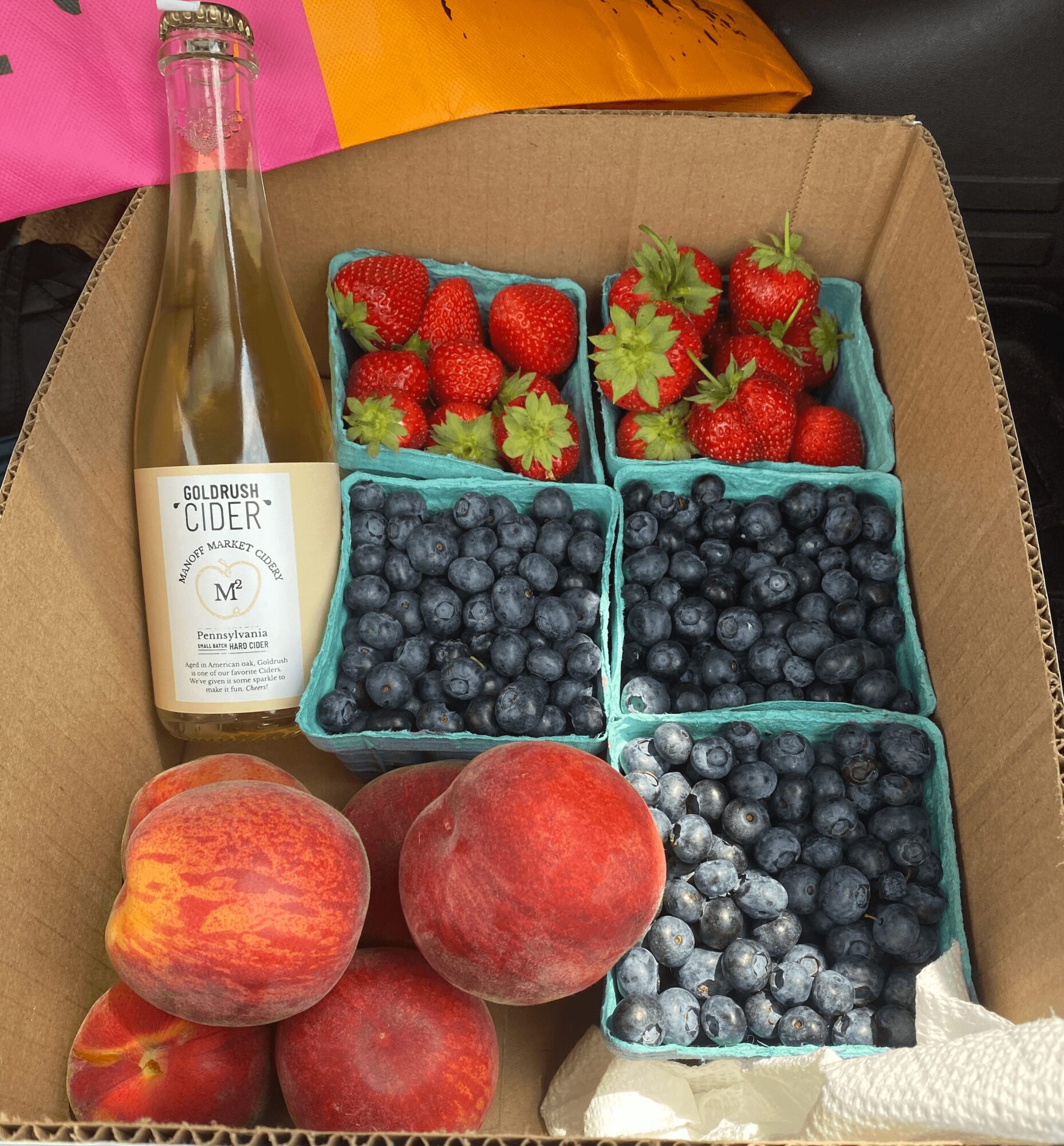 Box with strawberries, blueberries, peaches, a bottle, and paper towel, all arranged inside a cardboard box.