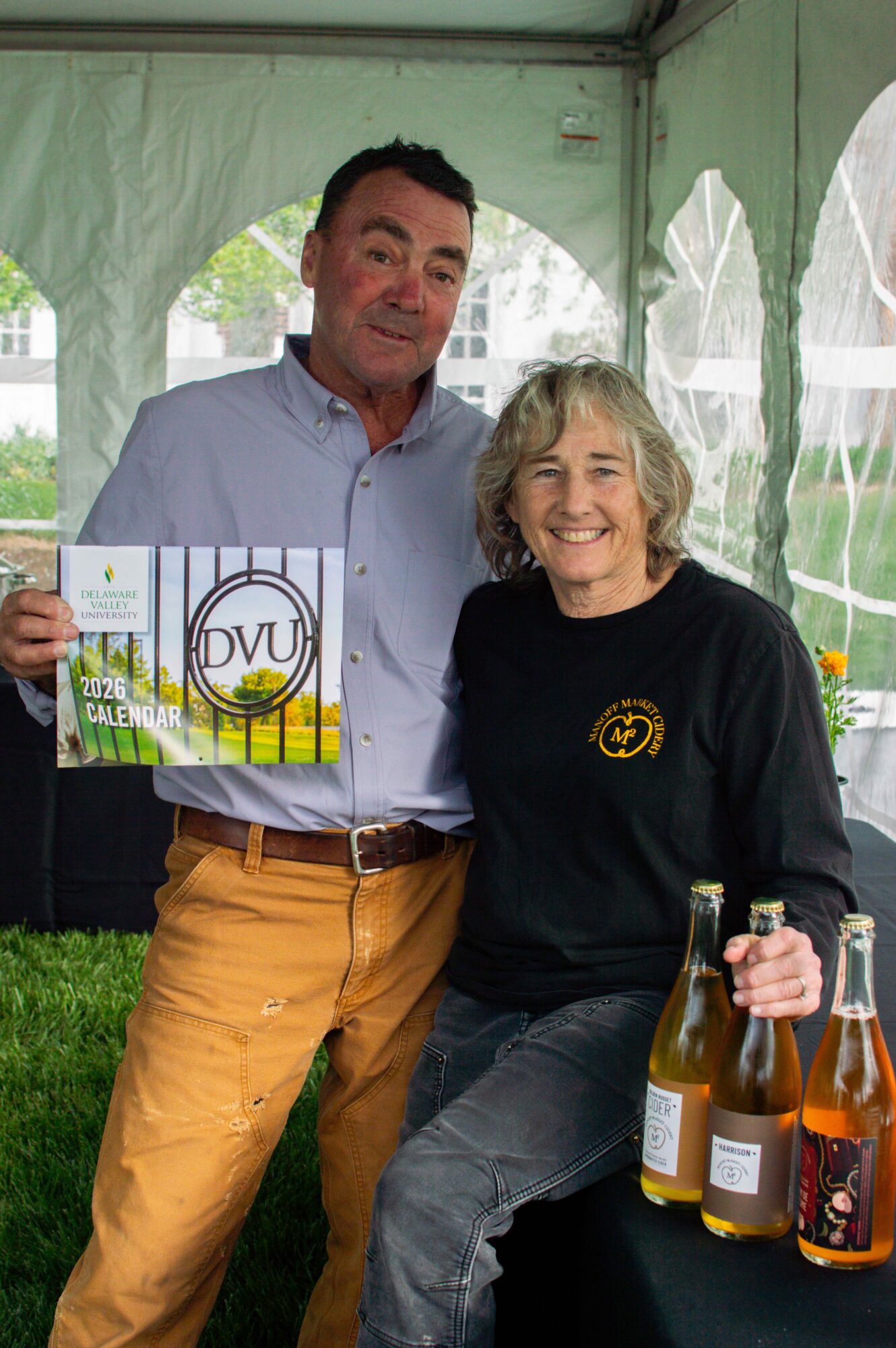 Two people smiling, one holding a magazine, inside a tent with bottles on a table, outdoors.