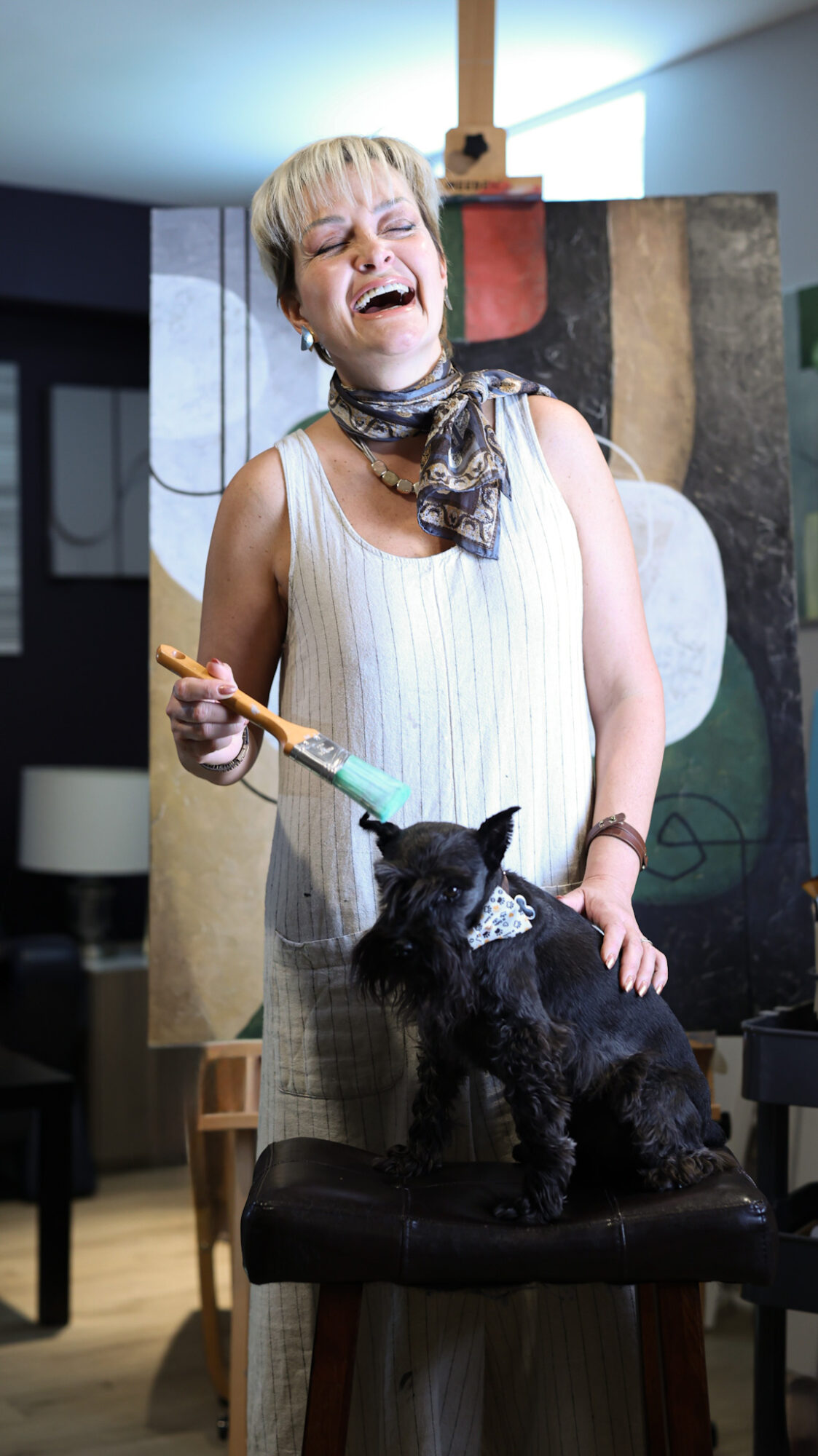Woman laughing with a small black dog on a chair, holding a toy, in a room with colorful artwork in the background.