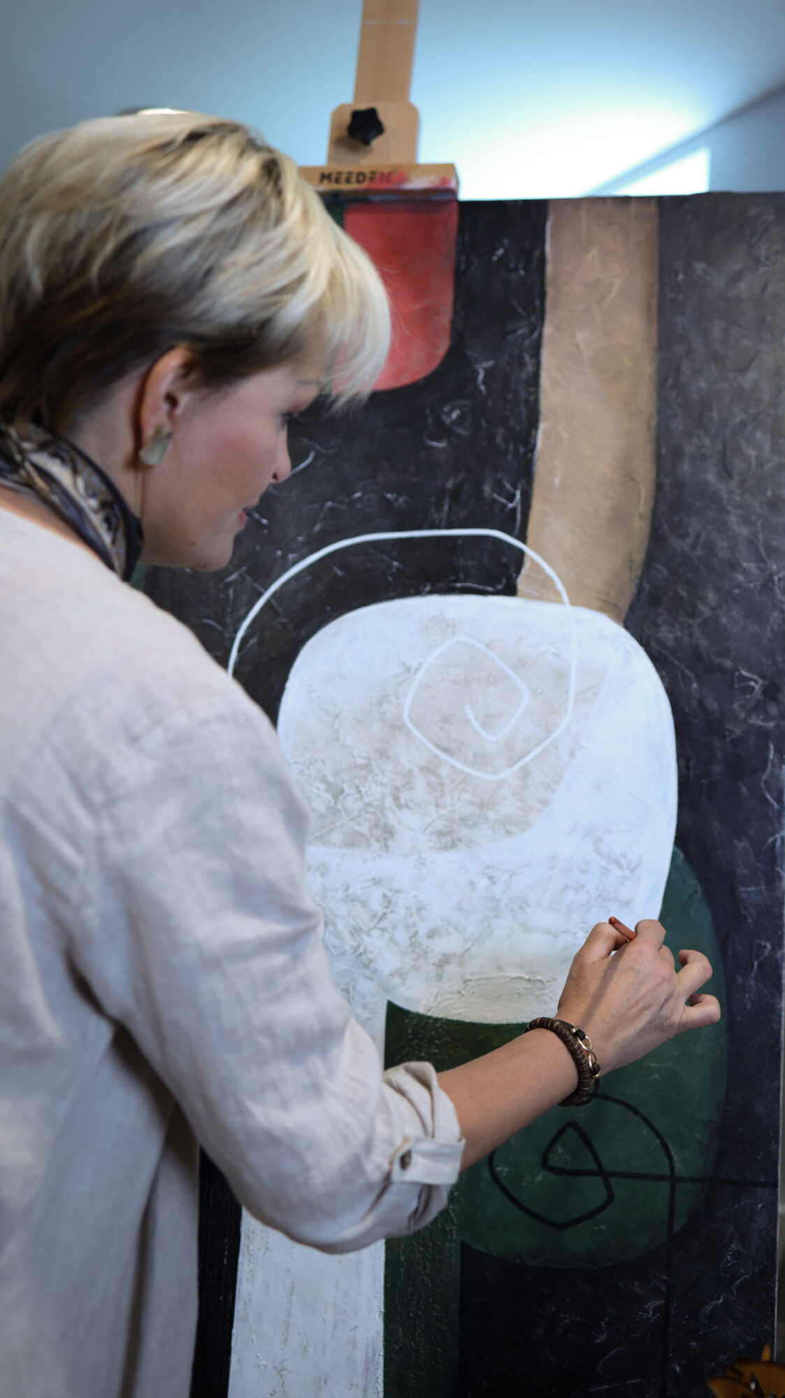 Person with short blonde hair drawing on a large canvas with abstract shapes and colors.