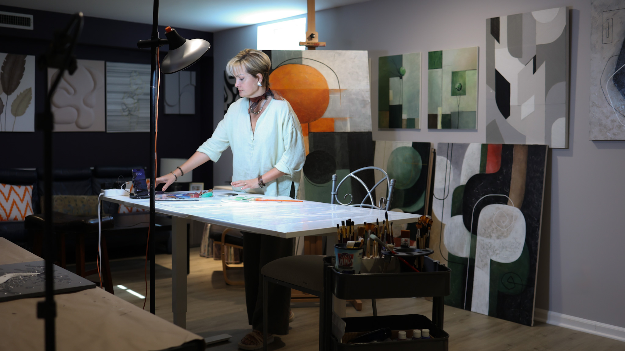 Person working at a table in an art studio with paintings on the wall behind them.