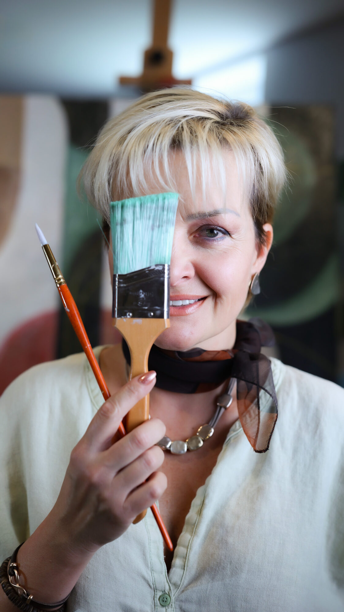 Woman holding a paintbrush and a palette with paint, smiling, in an indoor setting.
