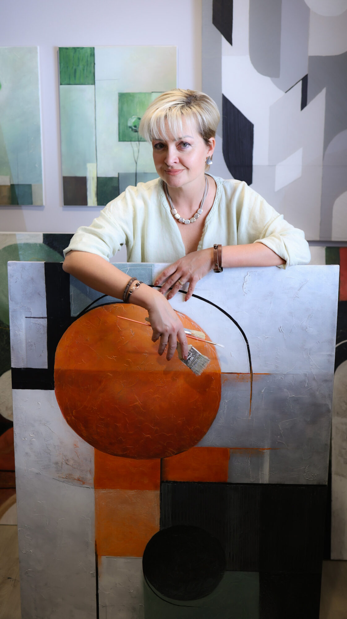 Woman with short blonde hair wearing a light-colored top, standing behind an abstract painting with geometric shapes and colors.
