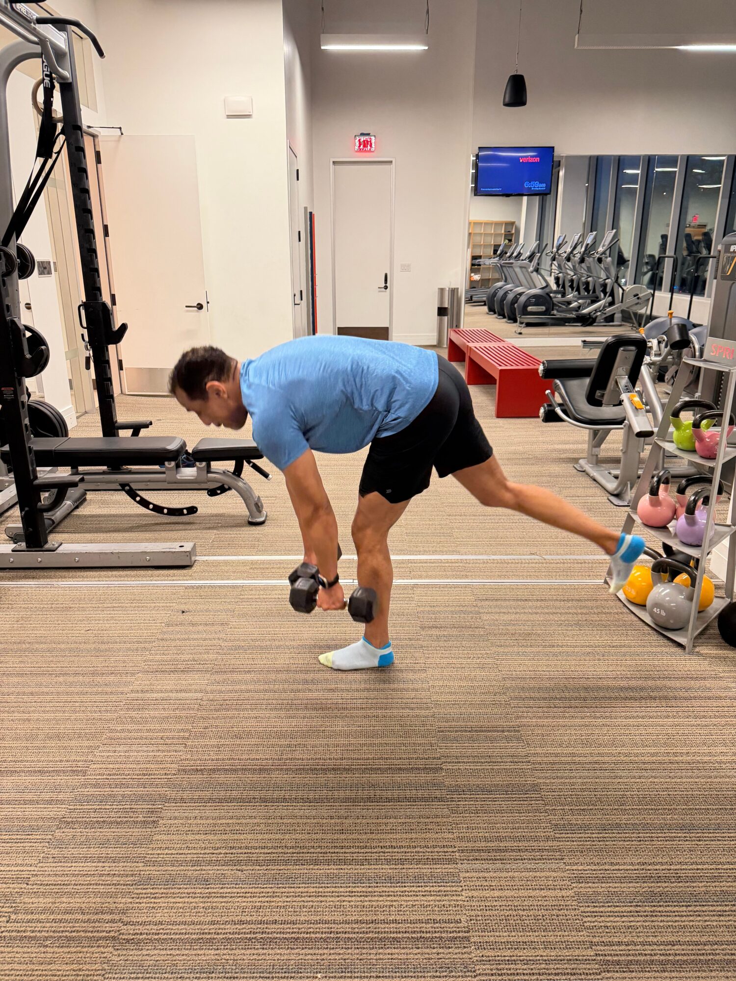Person stretching in a gym, holding a dumbbell, with gym equipment and a mirror in the background.