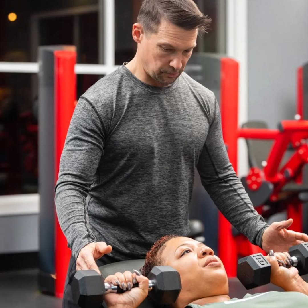 Man spotting woman lifting dumbbells in gym, both focused on workout, red gym equipment in background.