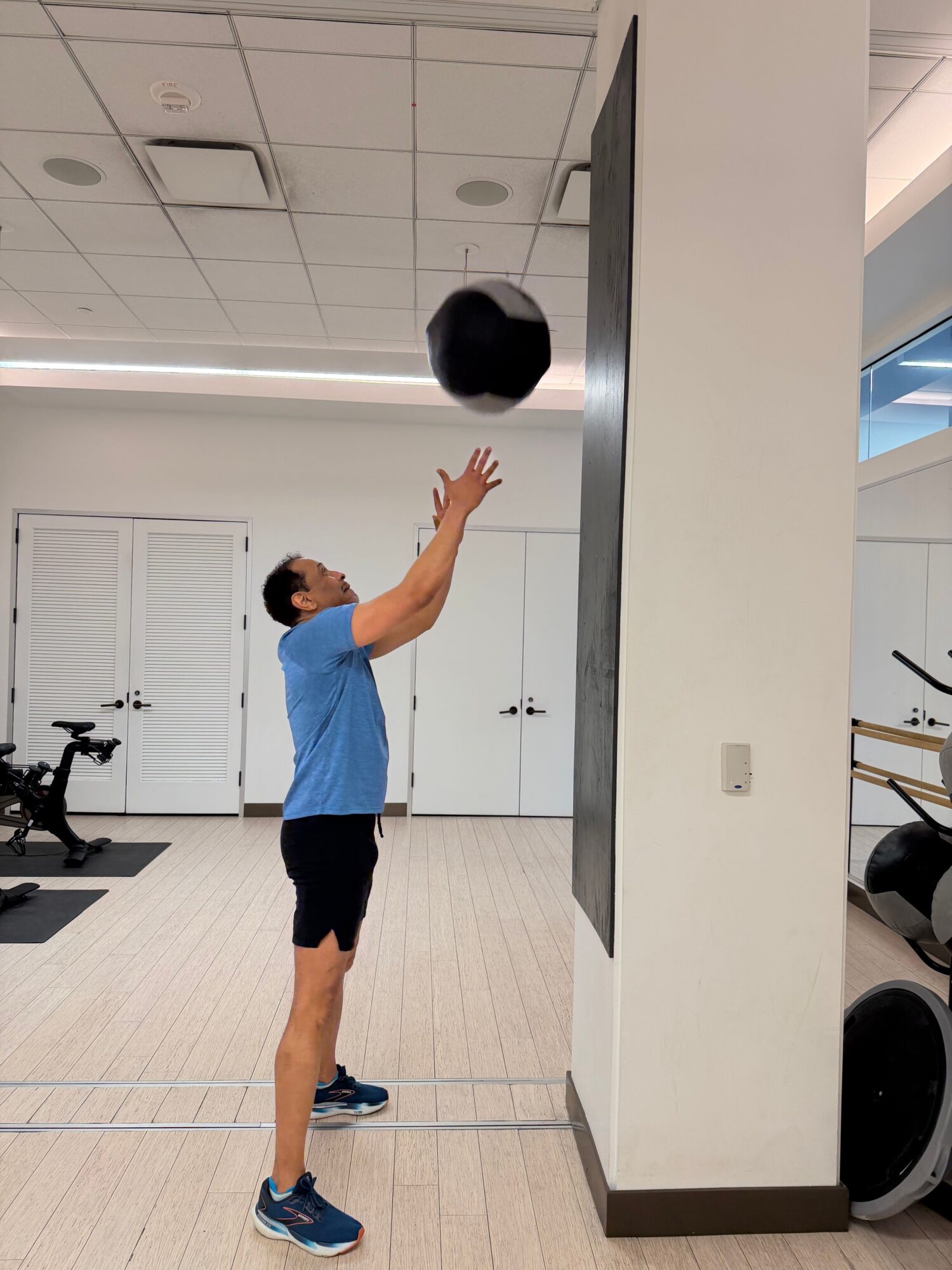 Person stretching arm in a room with white walls and tiled floor, wearing blue shirt and black shorts.