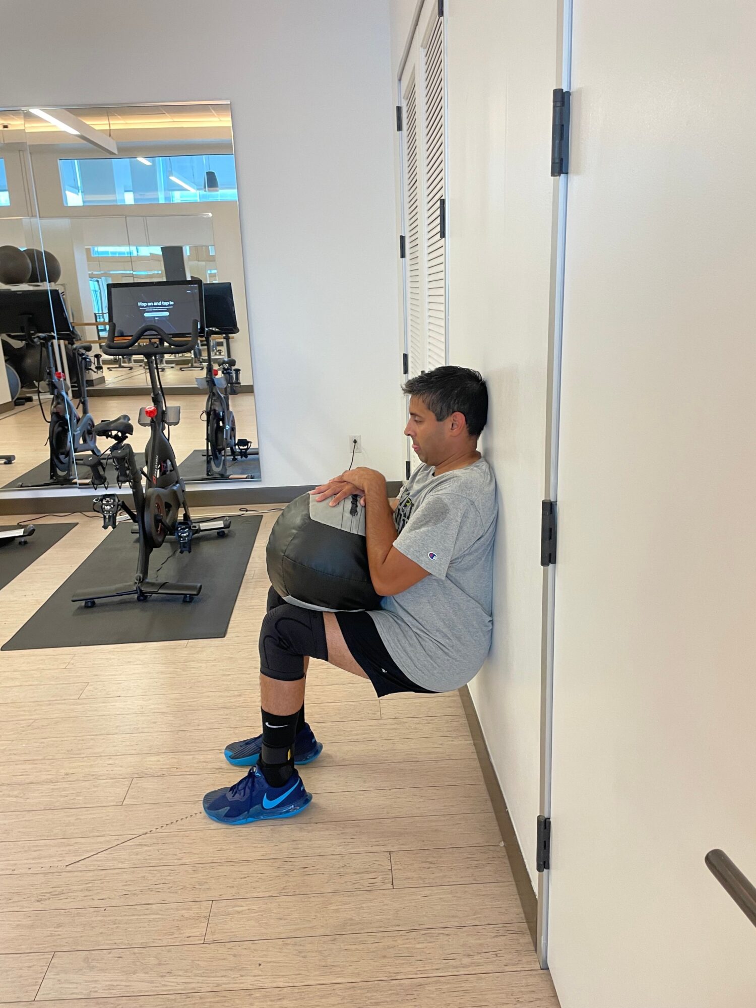 Person in a gym or fitness area, bending over a black exercise ball, with gym equipment and a mirror in the background.