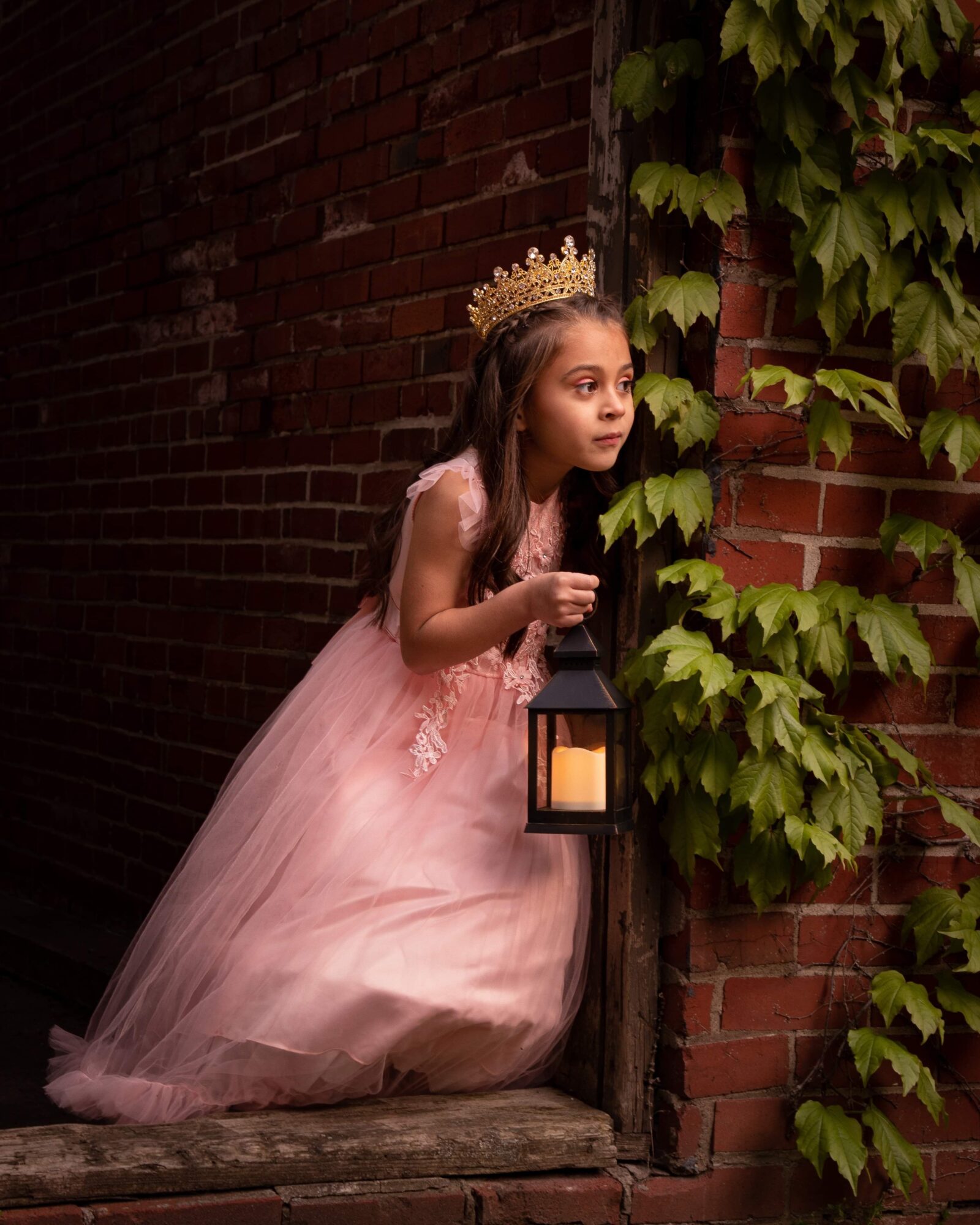 Young girl in a pink dress and crown looks through green ivy on a brick wall, holding a lantern.