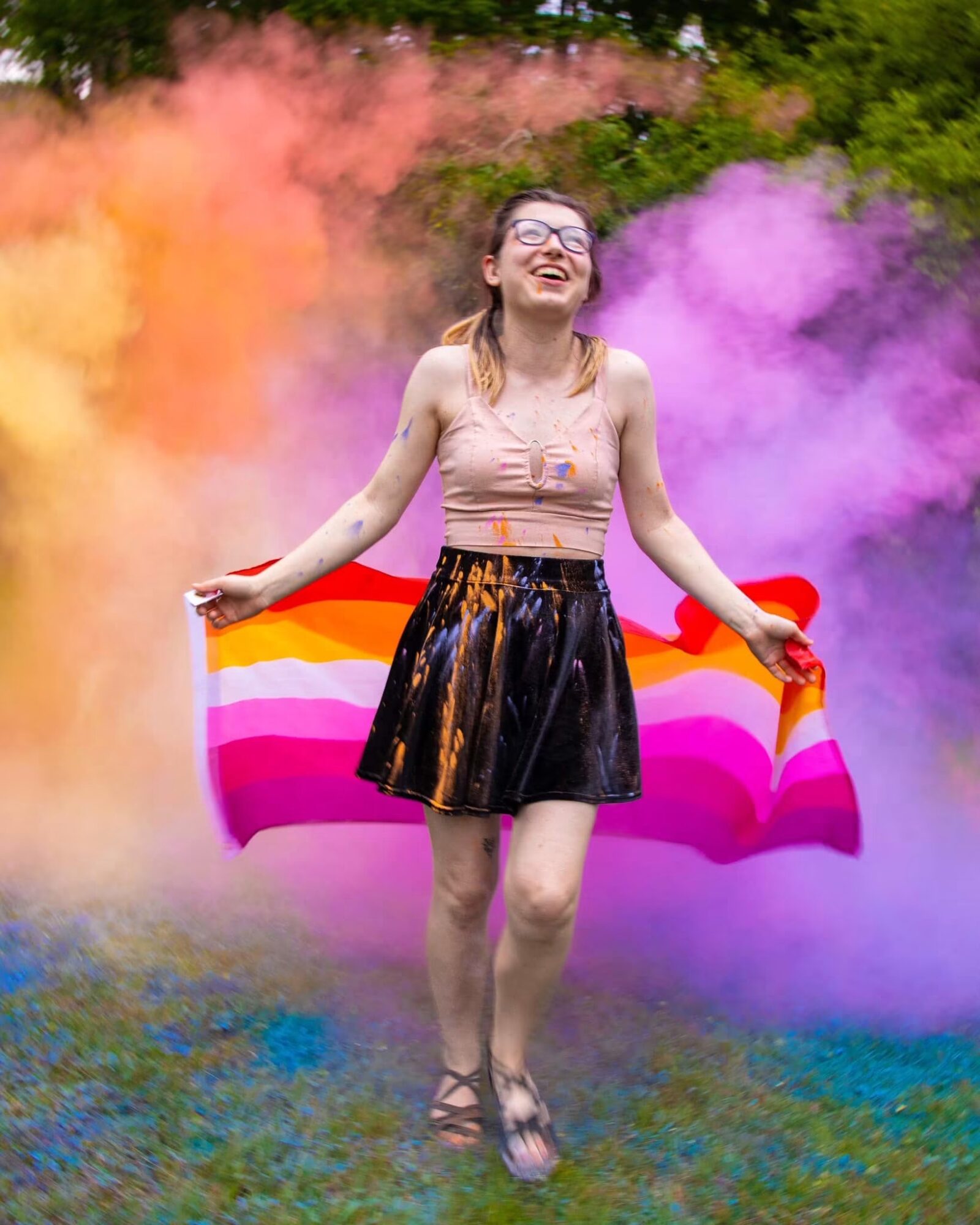 Young woman smiling, holding a rainbow pride flag, standing outdoors with colorful smoke in the background.