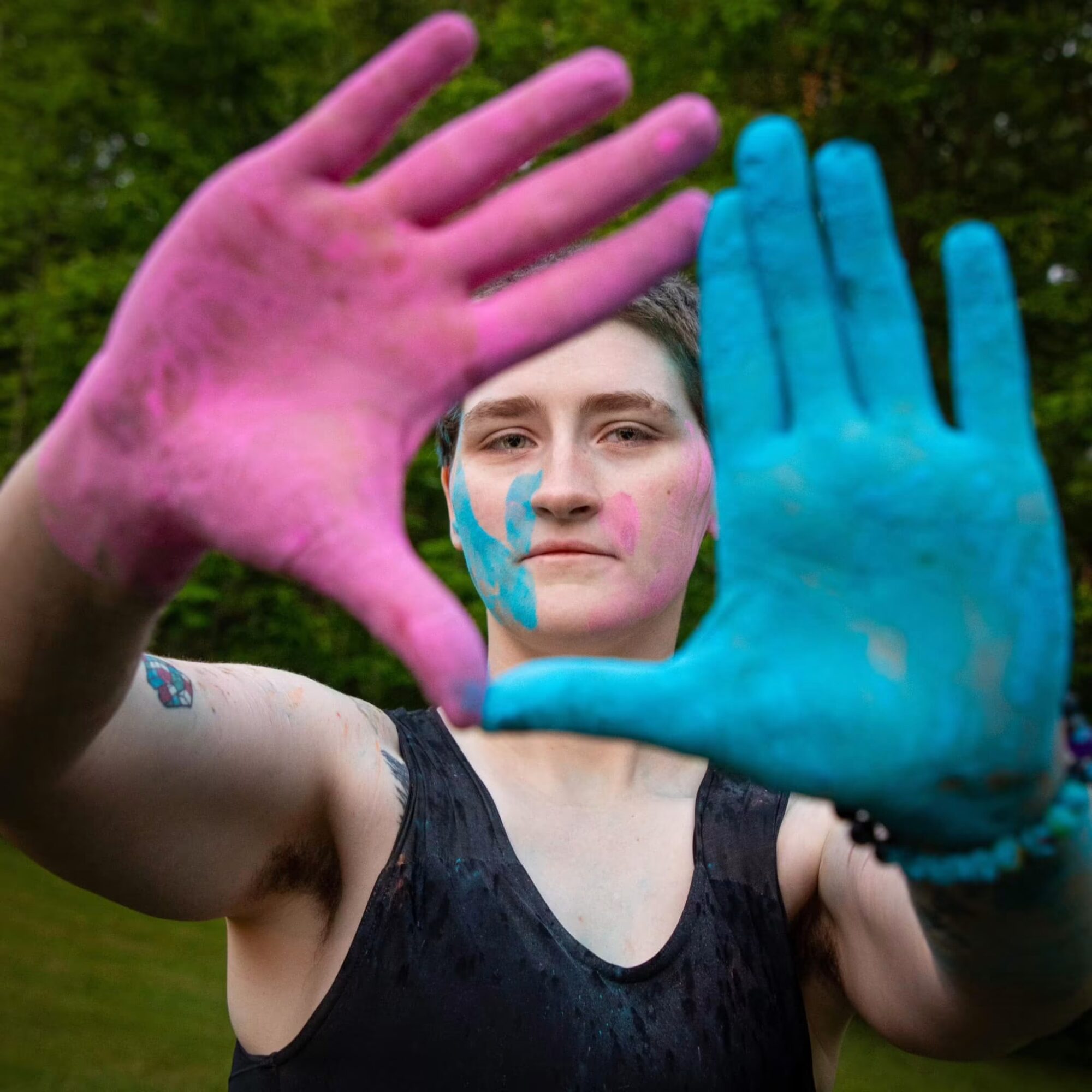 Person with painted hands holding up hands in front, outdoors with green background, wearing a black sleeveless top.