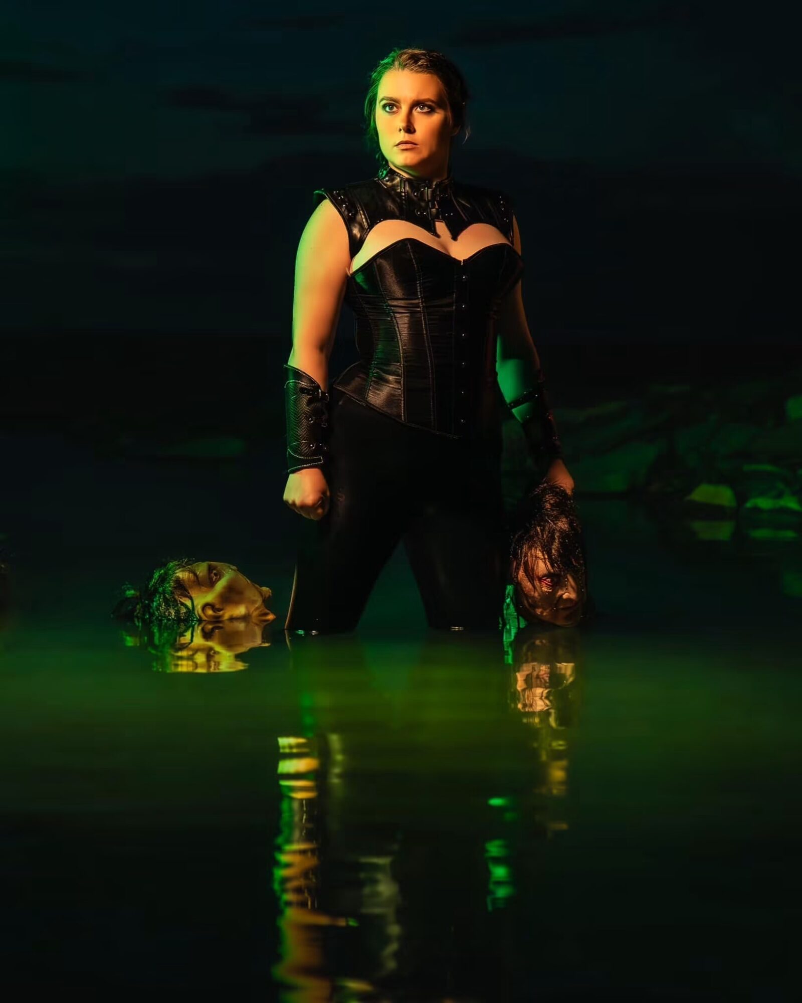 Woman in black outfit standing in water at night, with a skull and a mask nearby, illuminated by green light.