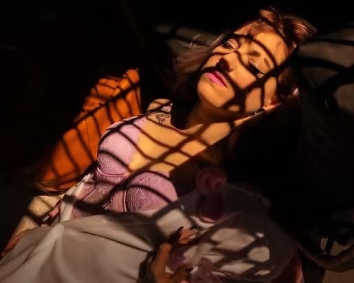 Woman lying down with shadows cast on her face and body, partially illuminated, with a pink top and dark background.