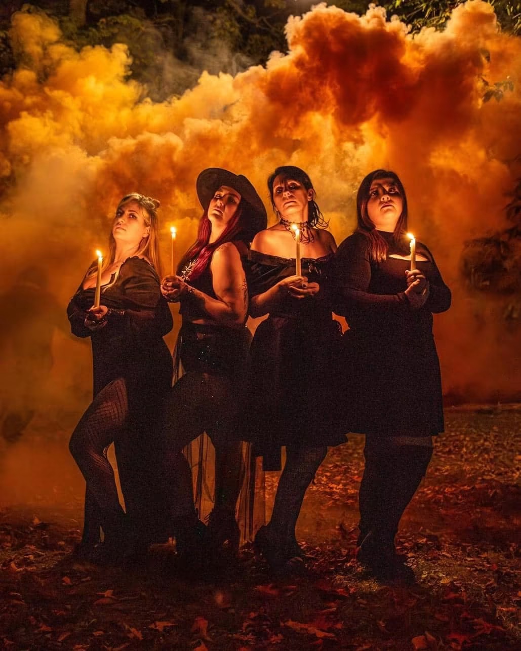 Four women in dark clothing holding candles stand outdoors at night with orange smoke in the background.