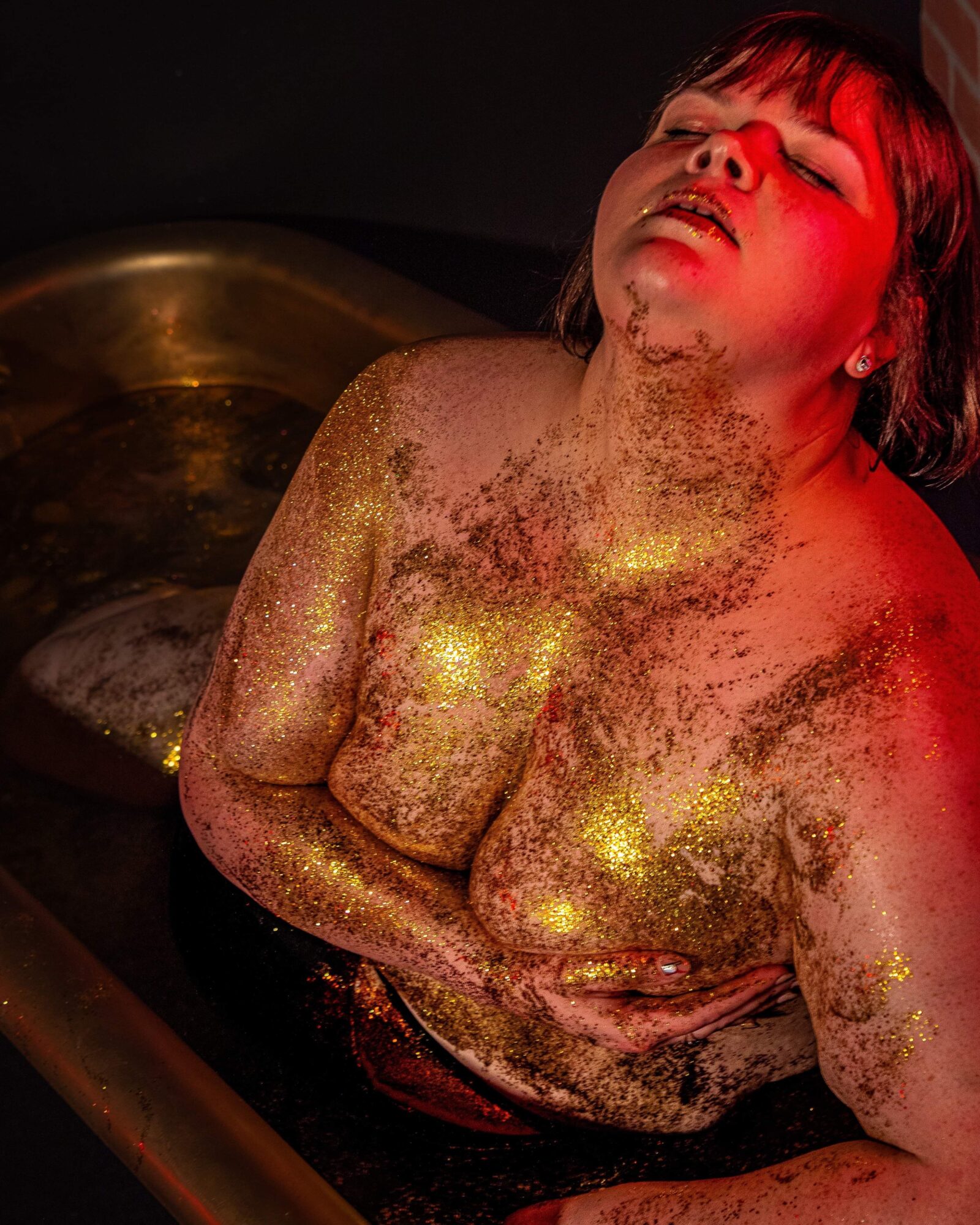 Person with short hair and closed eyes, covered in gold glitter, leaning back with a dark background.