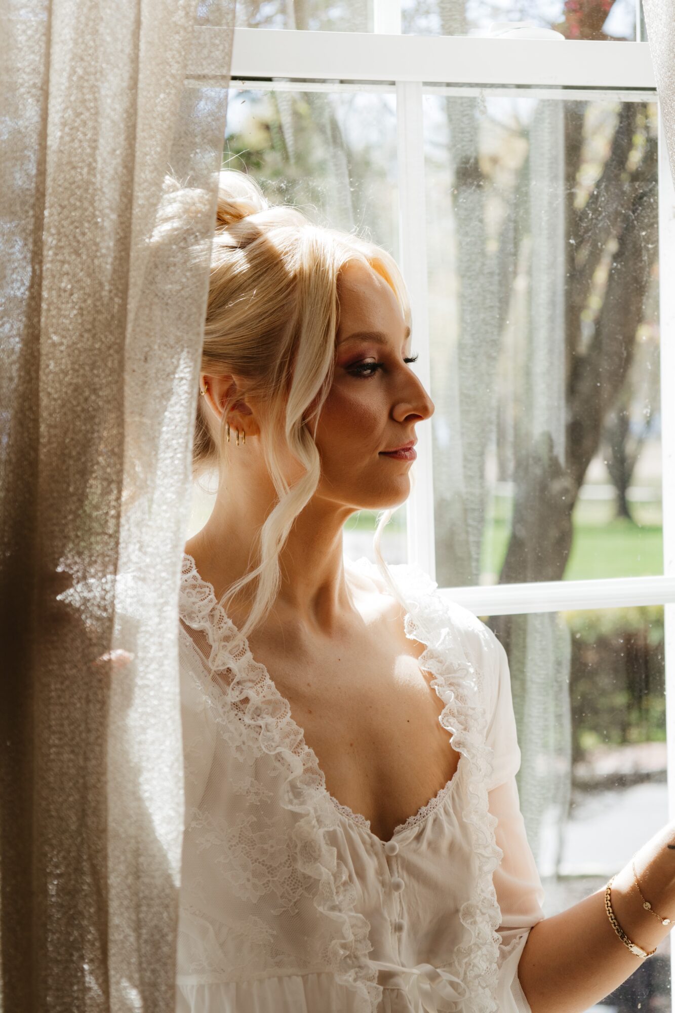 A woman with blonde hair in a loose updo, wearing a lace dress, looks out a window with sunlight streaming in.