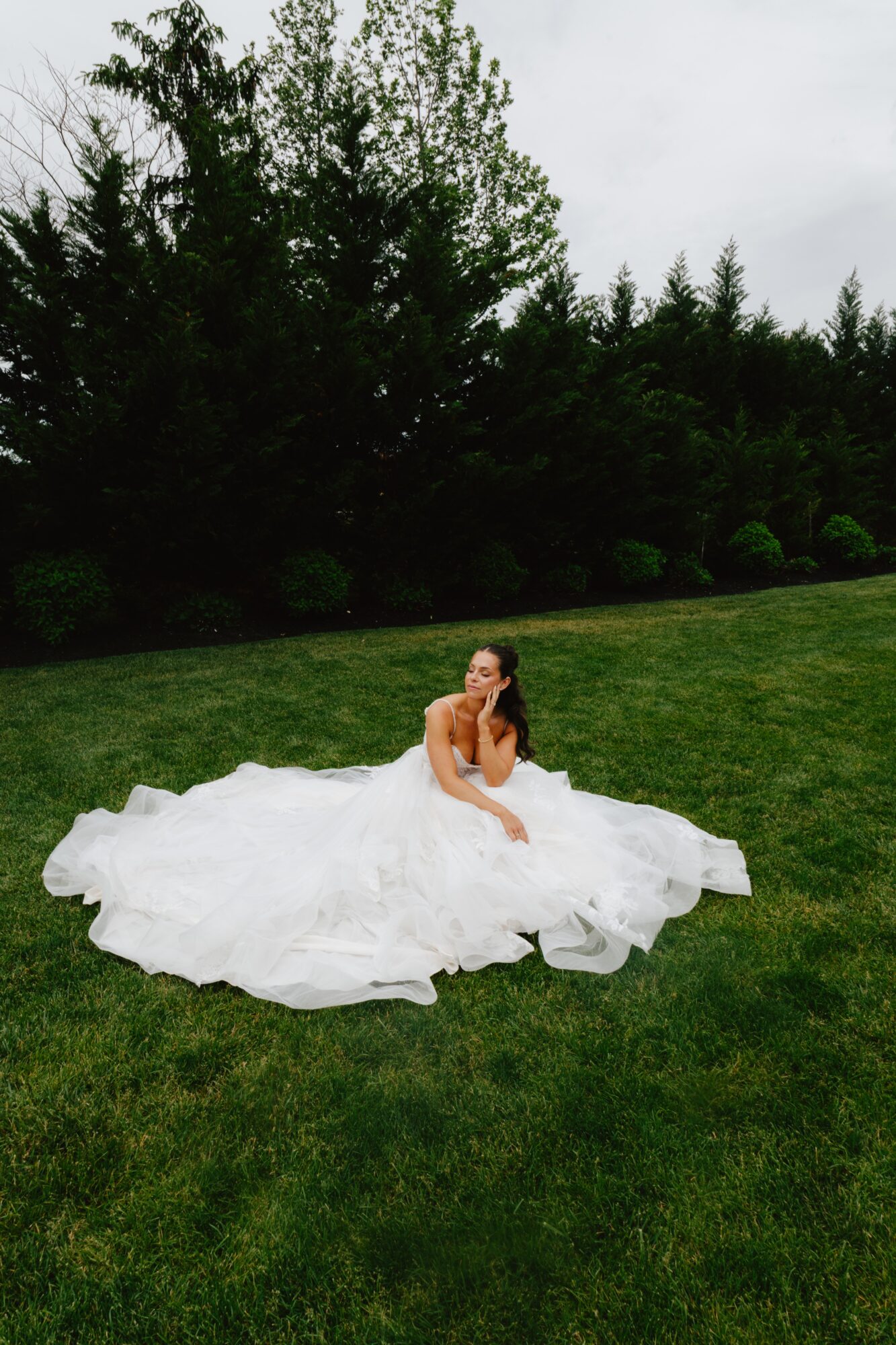 Bride in a white wedding dress sitting on grass, surrounded by trees and bushes, overcast sky, holding a bouquet.