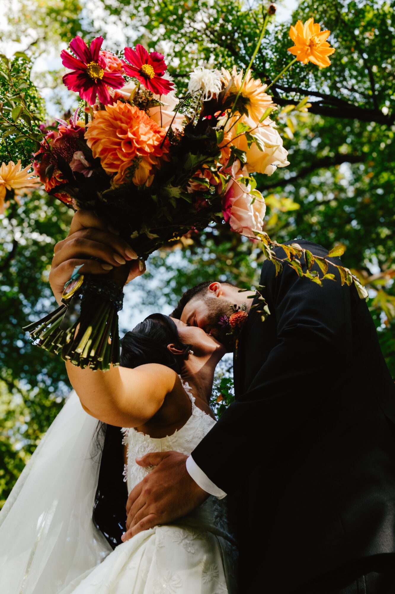 A couple in wedding attire kissing outdoors with a large bouquet of colorful flowers above them.