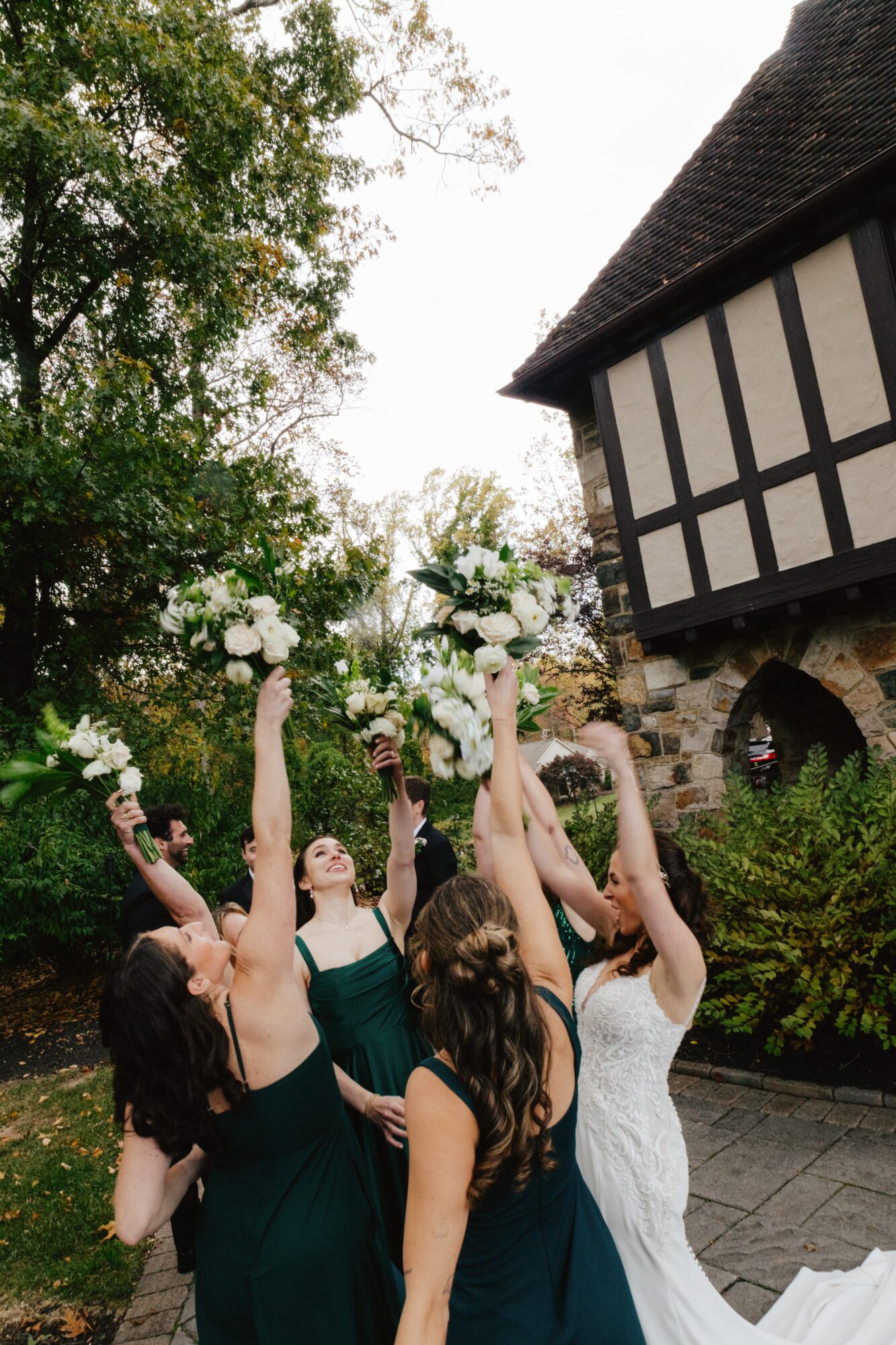 Group of women in formal dresses holding bouquets outdoors near a building with trees in the background.