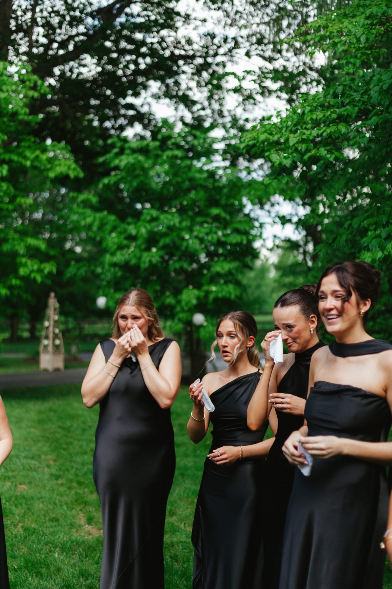 Four women in black dresses standing outdoors with green trees in the background.