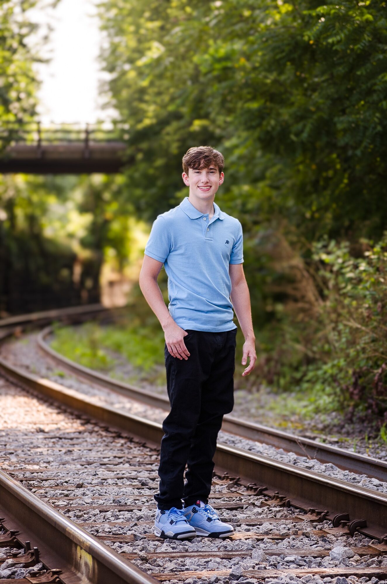 Young man standing on train tracks outdoors, smiling, with trees and a bridge in background.
