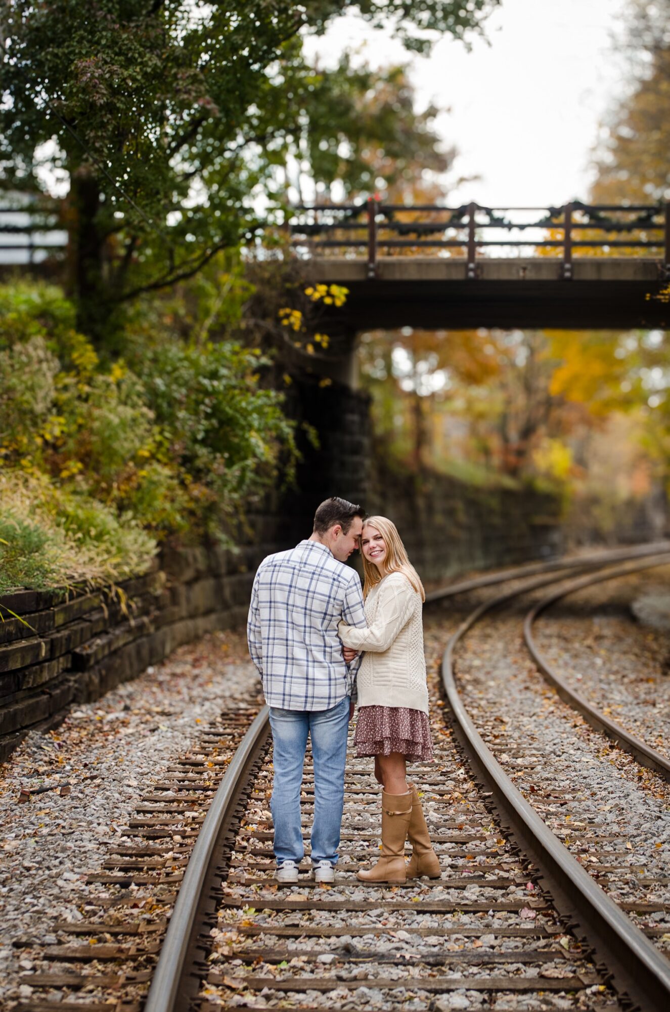 Couple standing on train tracks under a bridge, surrounded by trees with autumn leaves, embracing and looking at each other.