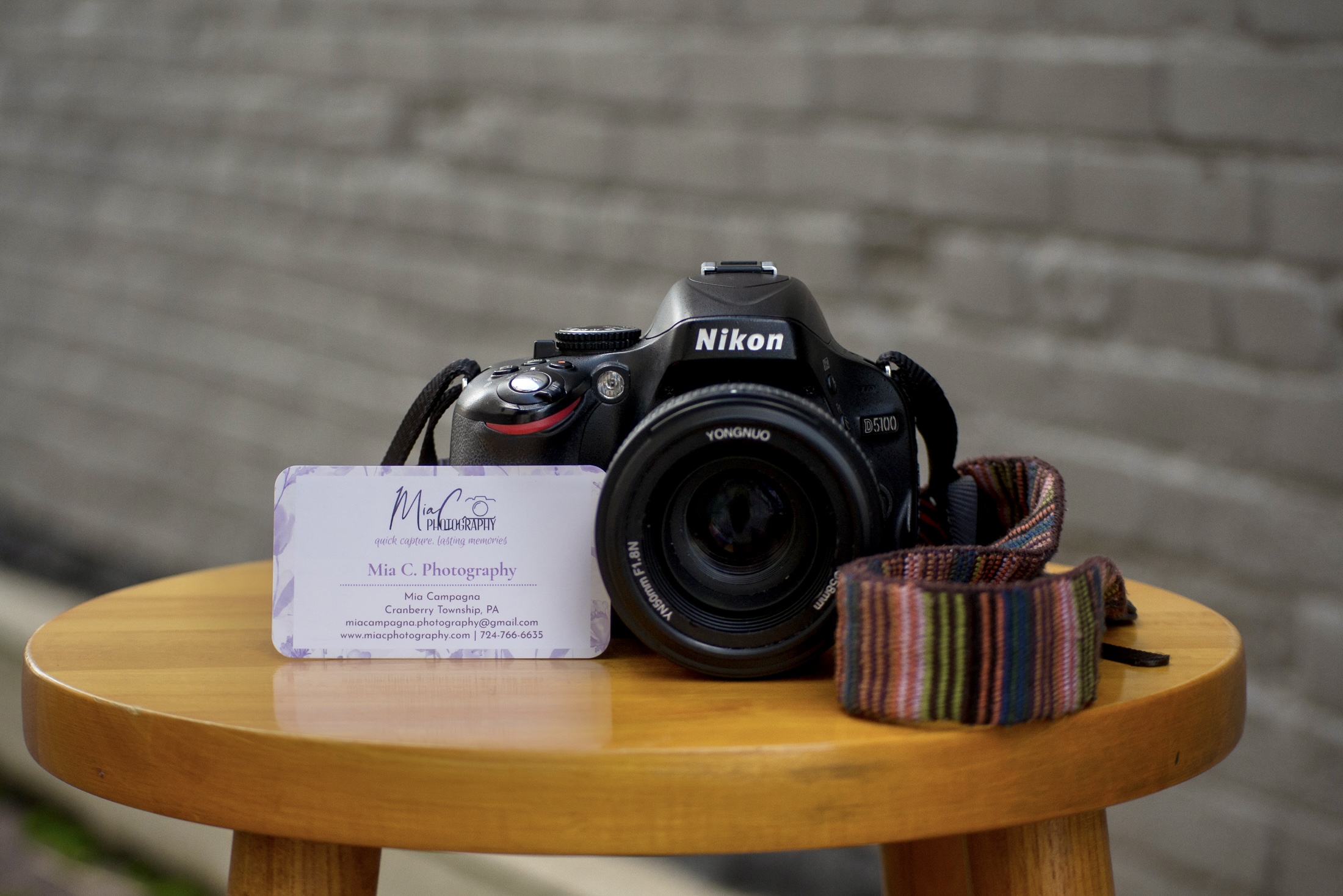 A Nikon DSLR camera with a colorful strap on a wooden table, with a business card in front, against a brick wall background.