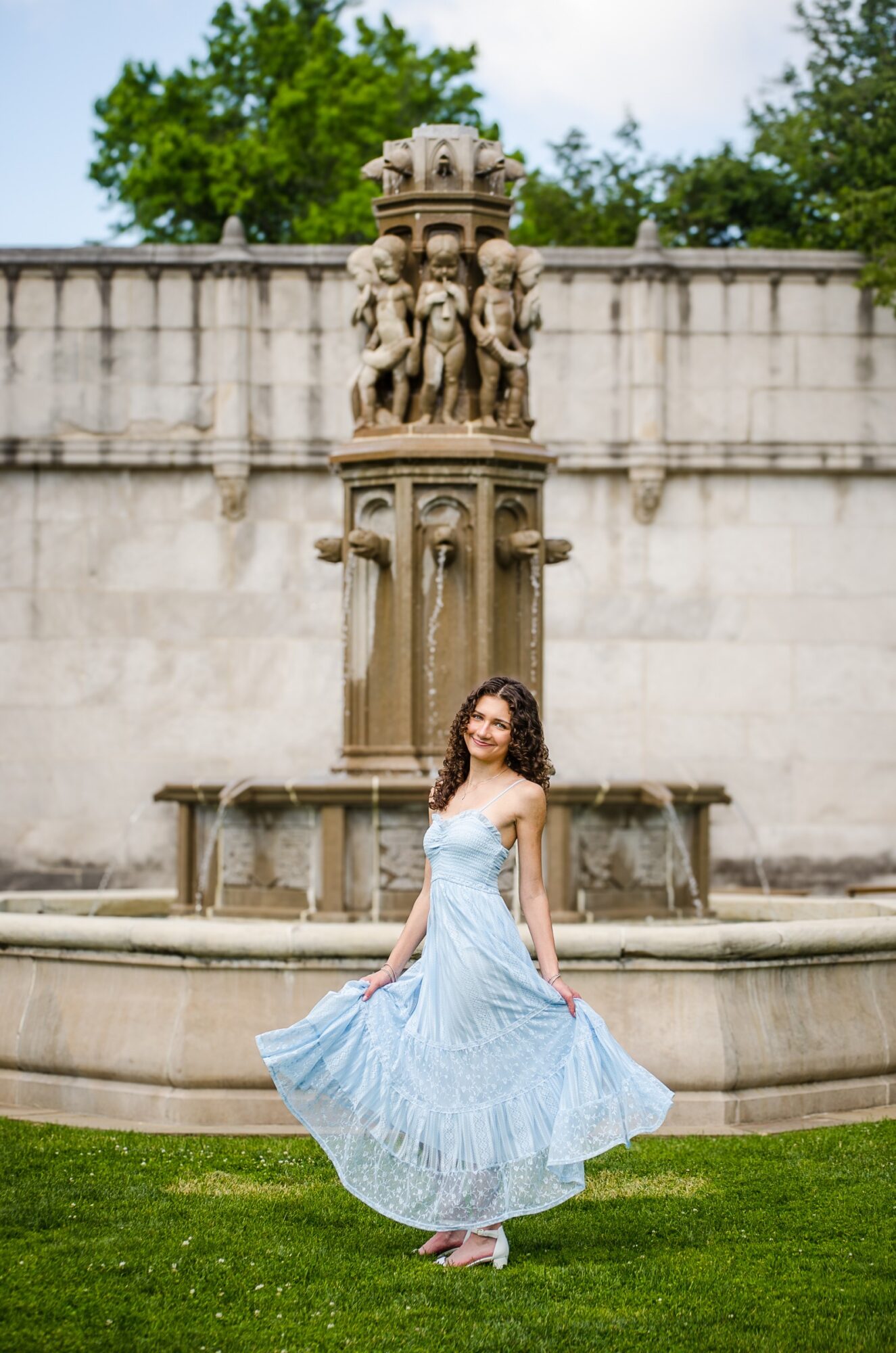 Woman in a light blue dress standing in front of a fountain with a stone sculpture, outdoors, green grass, trees, cloudy sky
