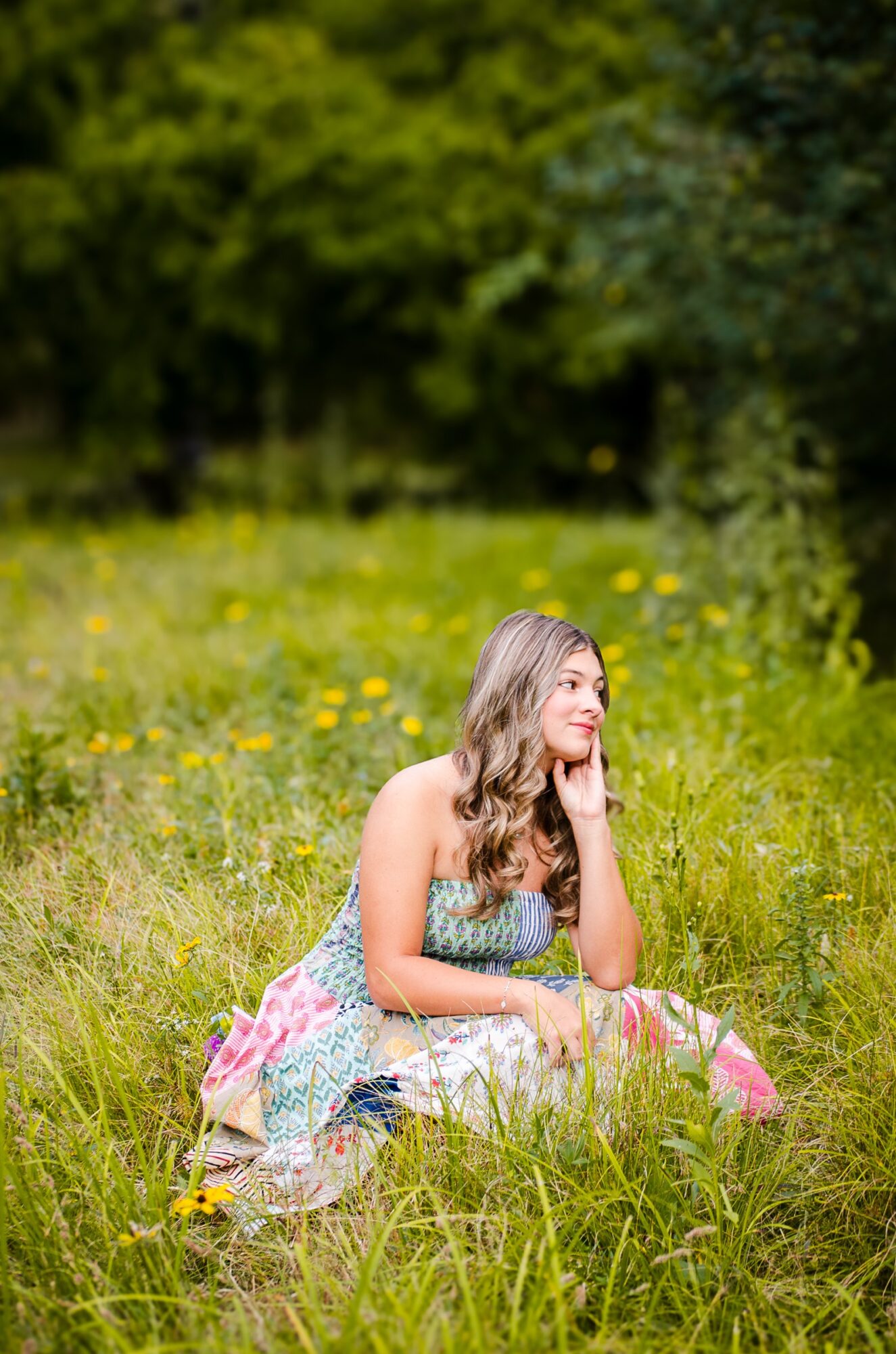 Young woman sitting in a grassy field with yellow flowers, surrounded by trees, resting her chin on her hand, looking to the side.