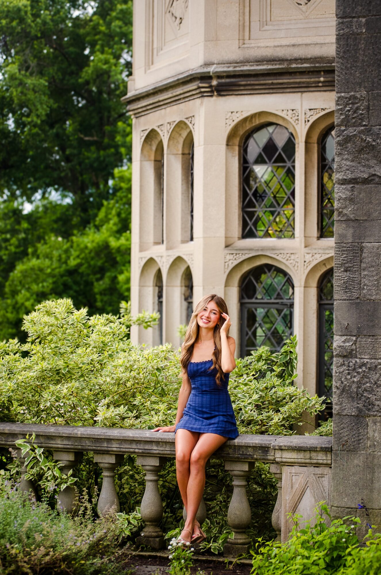 Young woman in blue dress sitting on stone railing in front of a historic building with arched windows, surrounded by greenery.