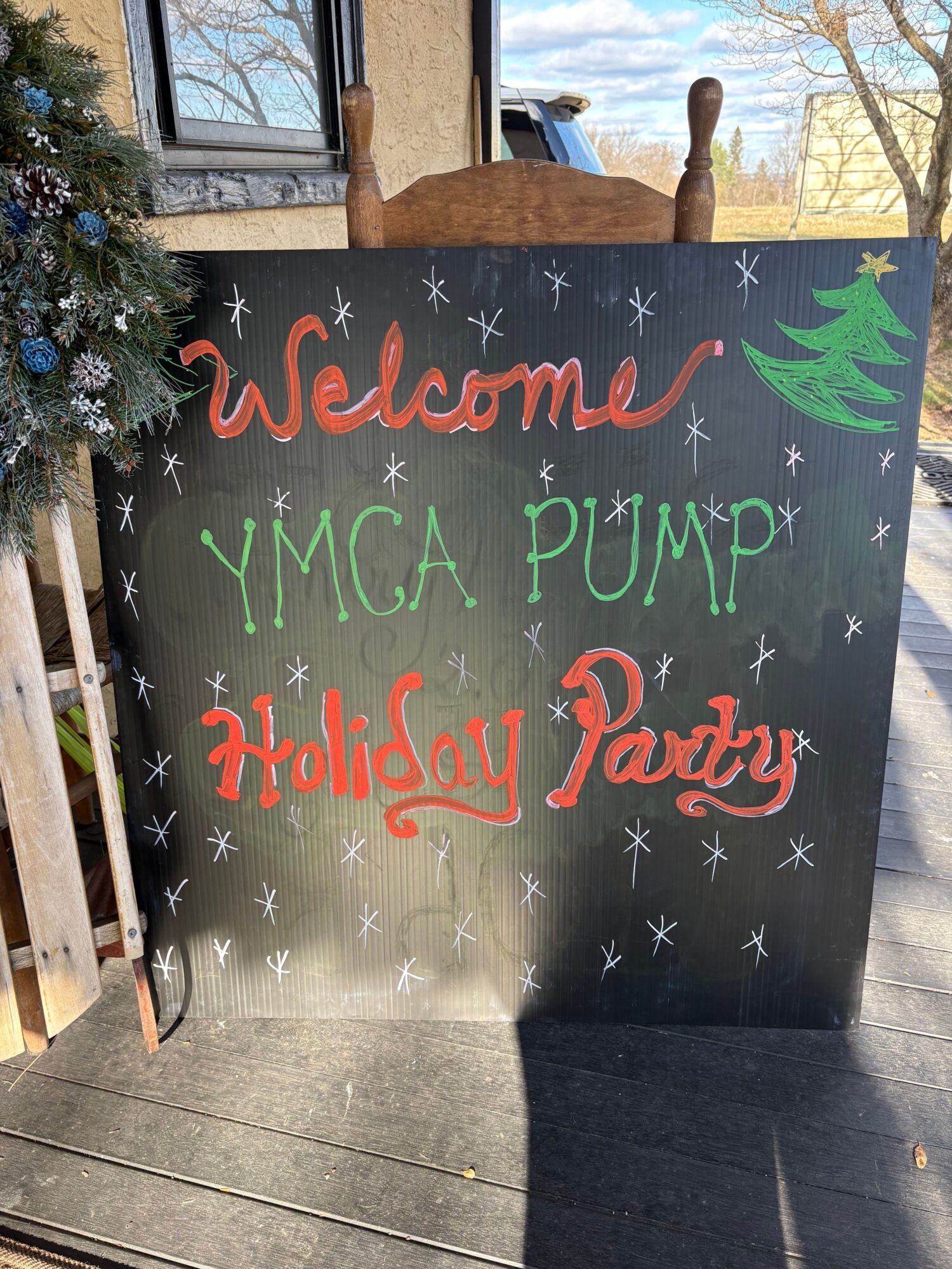 Blackboard with colorful handwritten text about summer, YMCA, and physical activity, decorated with stars and a tree, on a wooden deck.