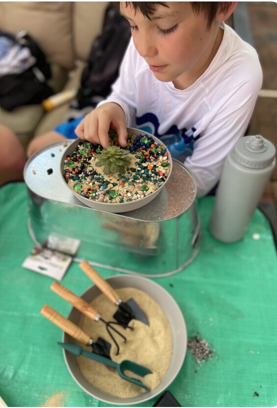 Child with short dark hair wearing a white shirt, working with a container of colorful sprinkles, a bowl of dough, and tools on a green table.