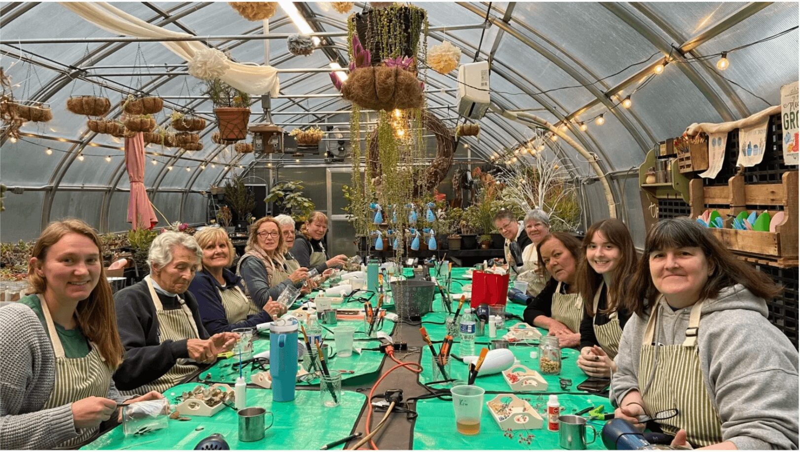 Group of people sitting at a long table inside a greenhouse, enjoying a meal or workshop, with plants and decorations overhead.