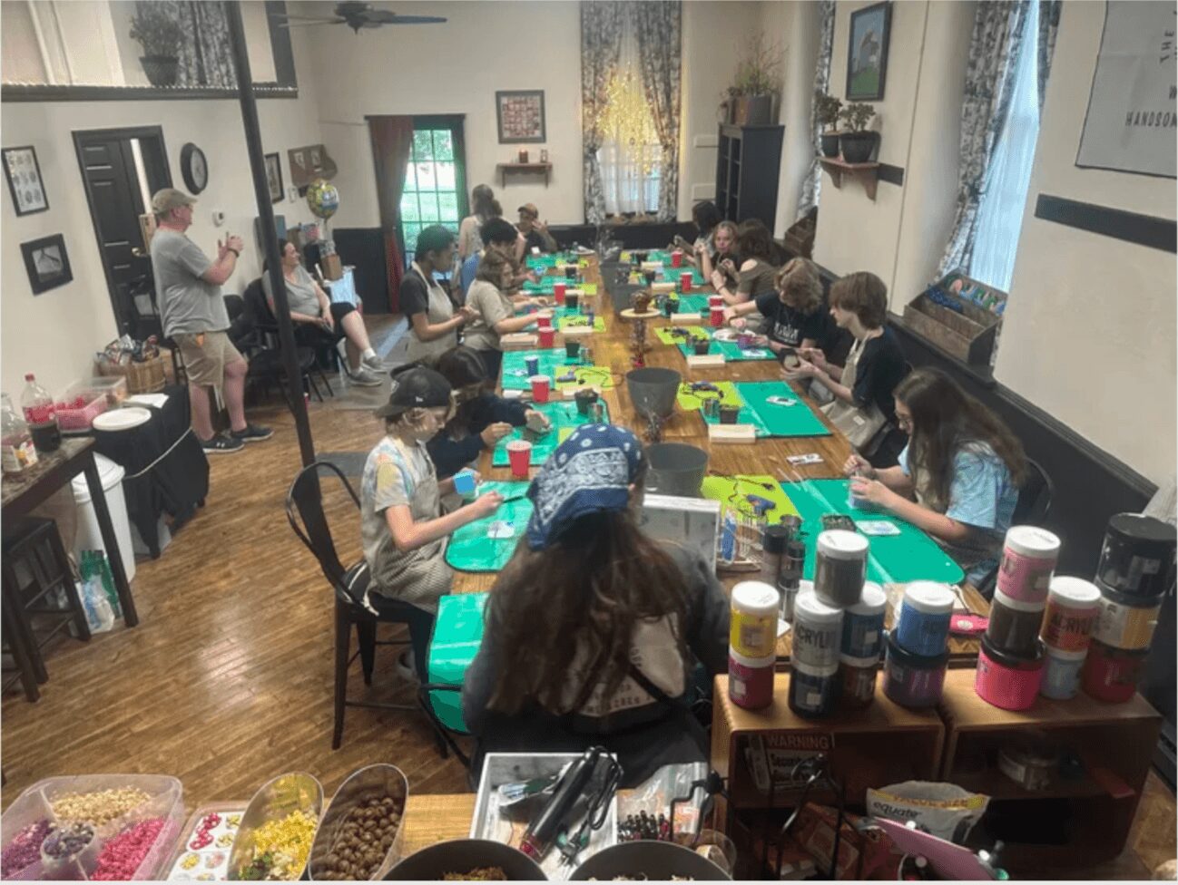 Children sit at a long table with art supplies, working on projects in a decorated room with windows and curtains.