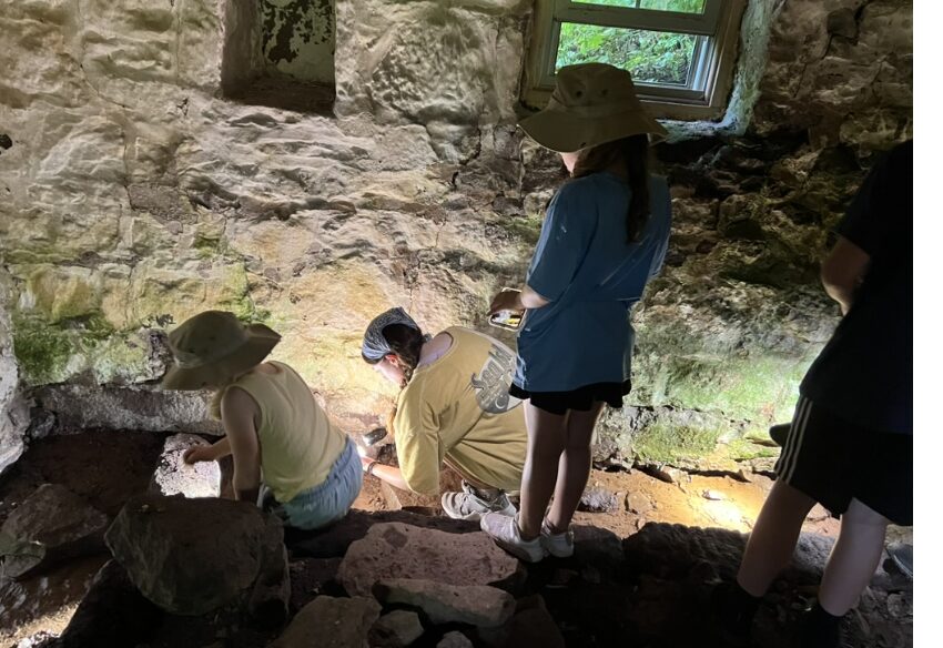 People exploring inside a rocky cave with a small window, some wearing hats and casual clothing, illuminated by natural light.
