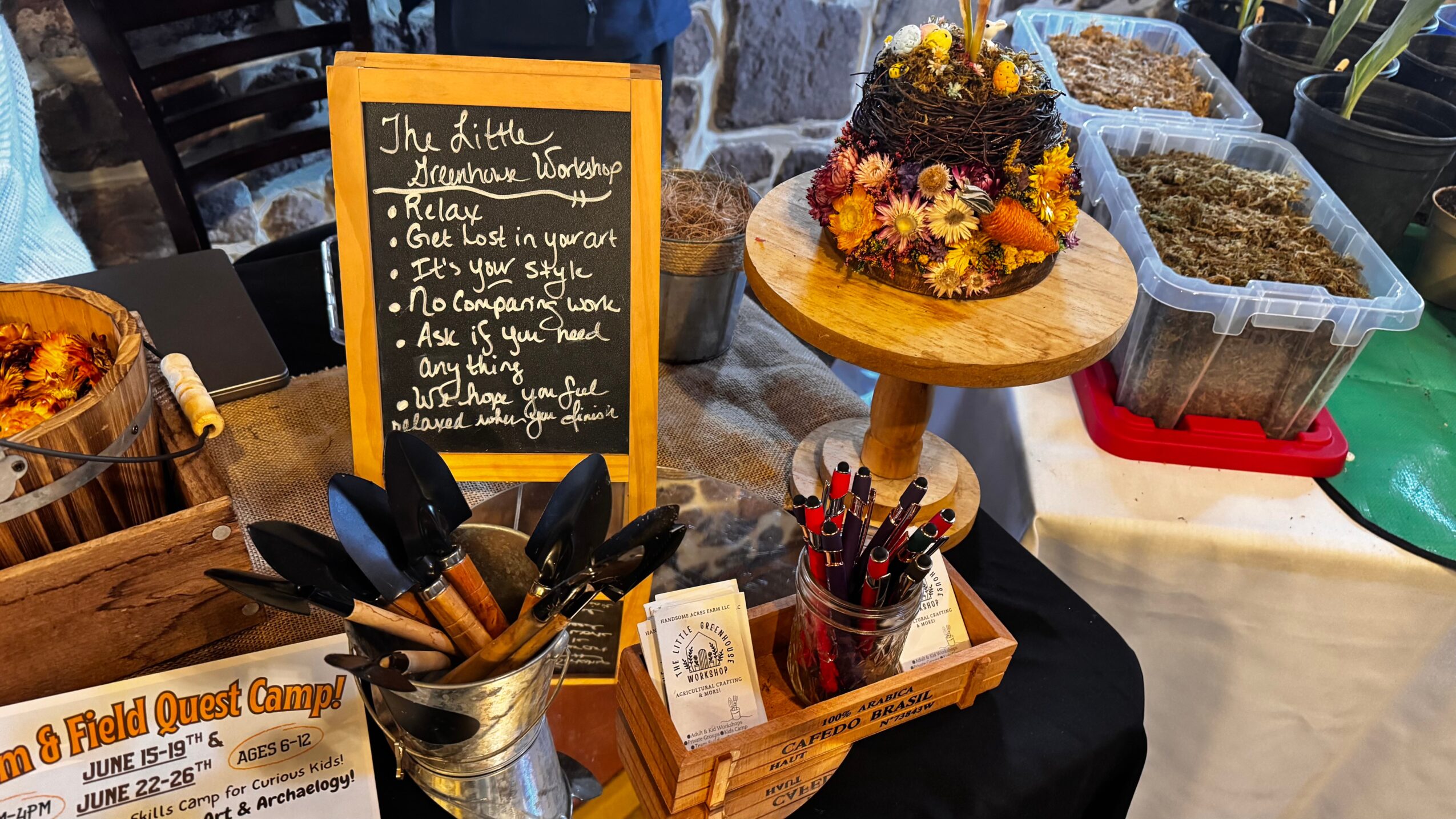 Display of a small cake decorated with flowers on a wooden stand, with containers of herbs or spices nearby, and a blackboard sign.