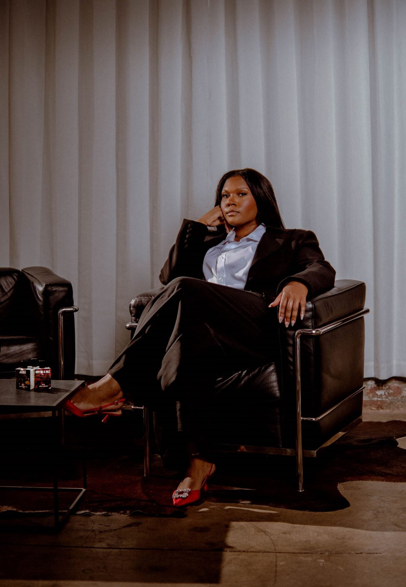 Woman sitting in a black armchair with a serious expression, wearing a dark blazer, white shirt, and red shoes, in front of a curtain.