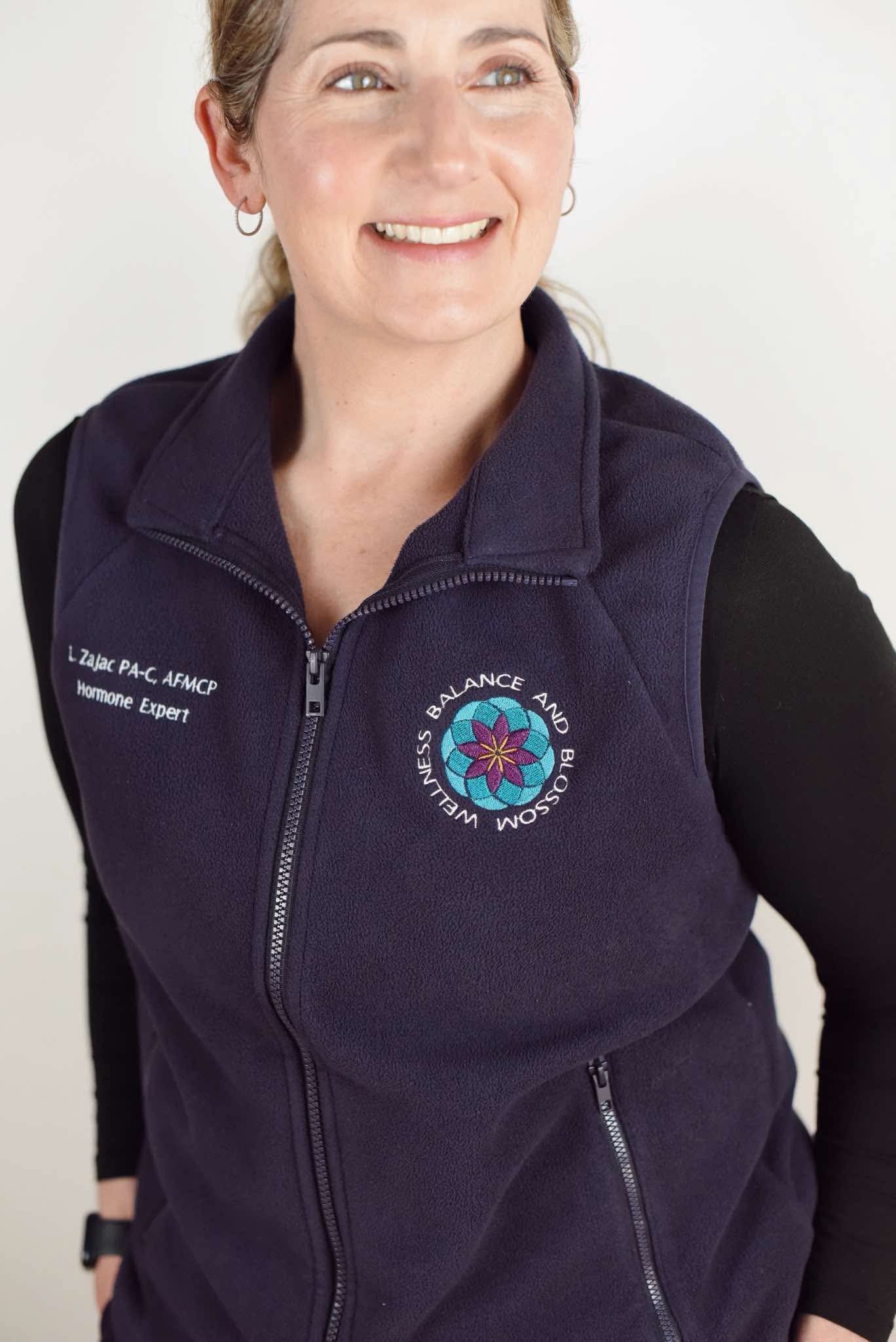 Woman smiling, wearing a navy vest with a logo on the chest, against a plain background.