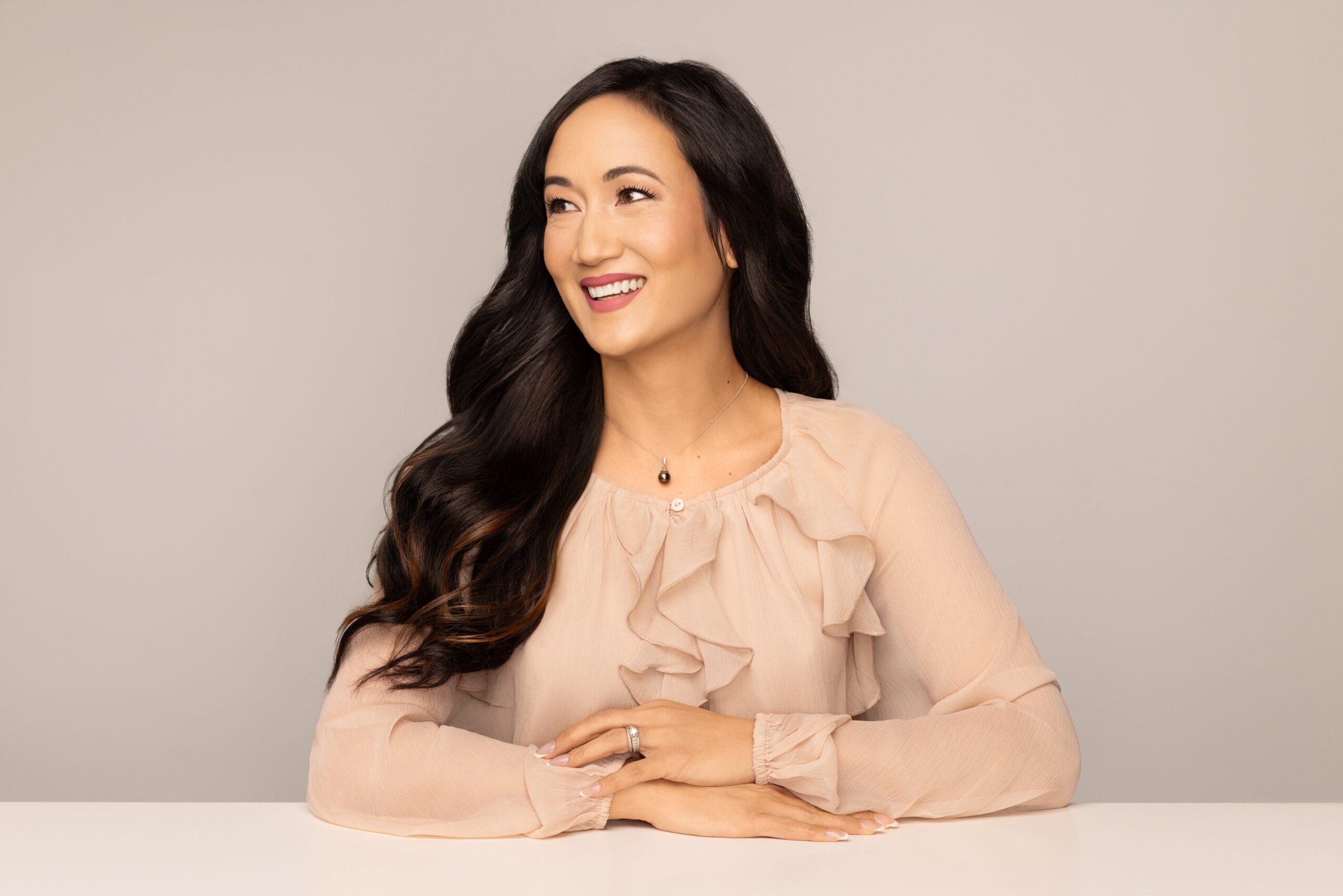 Woman with long dark hair smiling, wearing a beige blouse with ruffles, sitting at a table against a plain background.