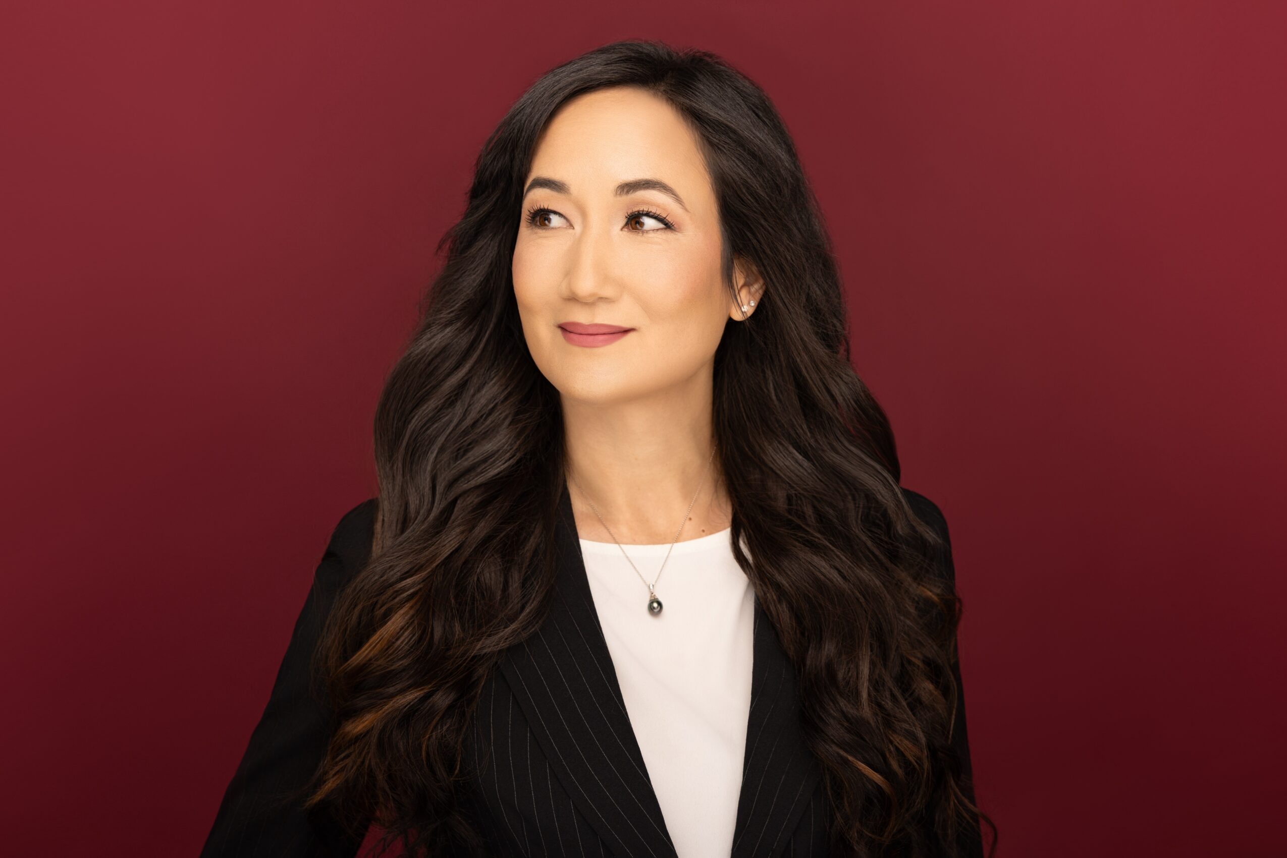 Woman with long wavy hair wearing a black blazer and white top, looking to the side, against a red background.