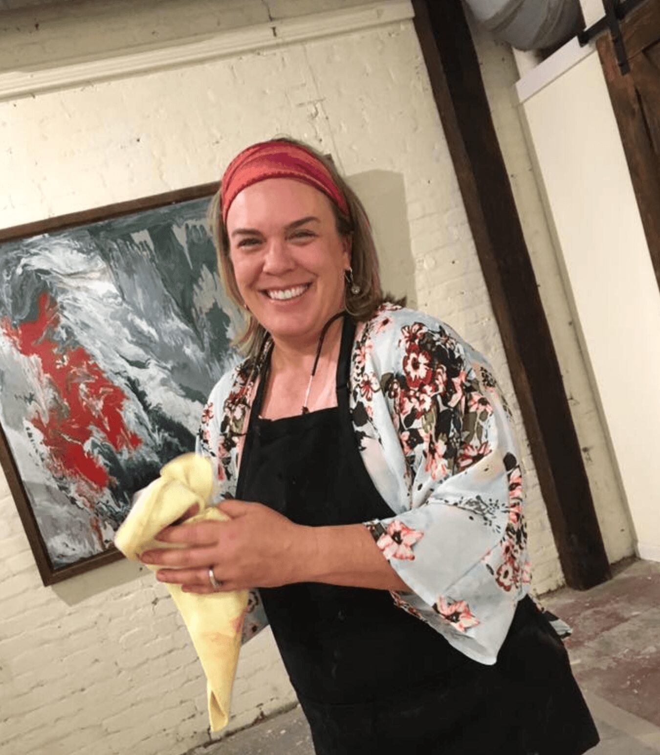 Woman with pink headband smiling, holding a cone-shaped pastry, standing indoors near a colorful abstract painting.