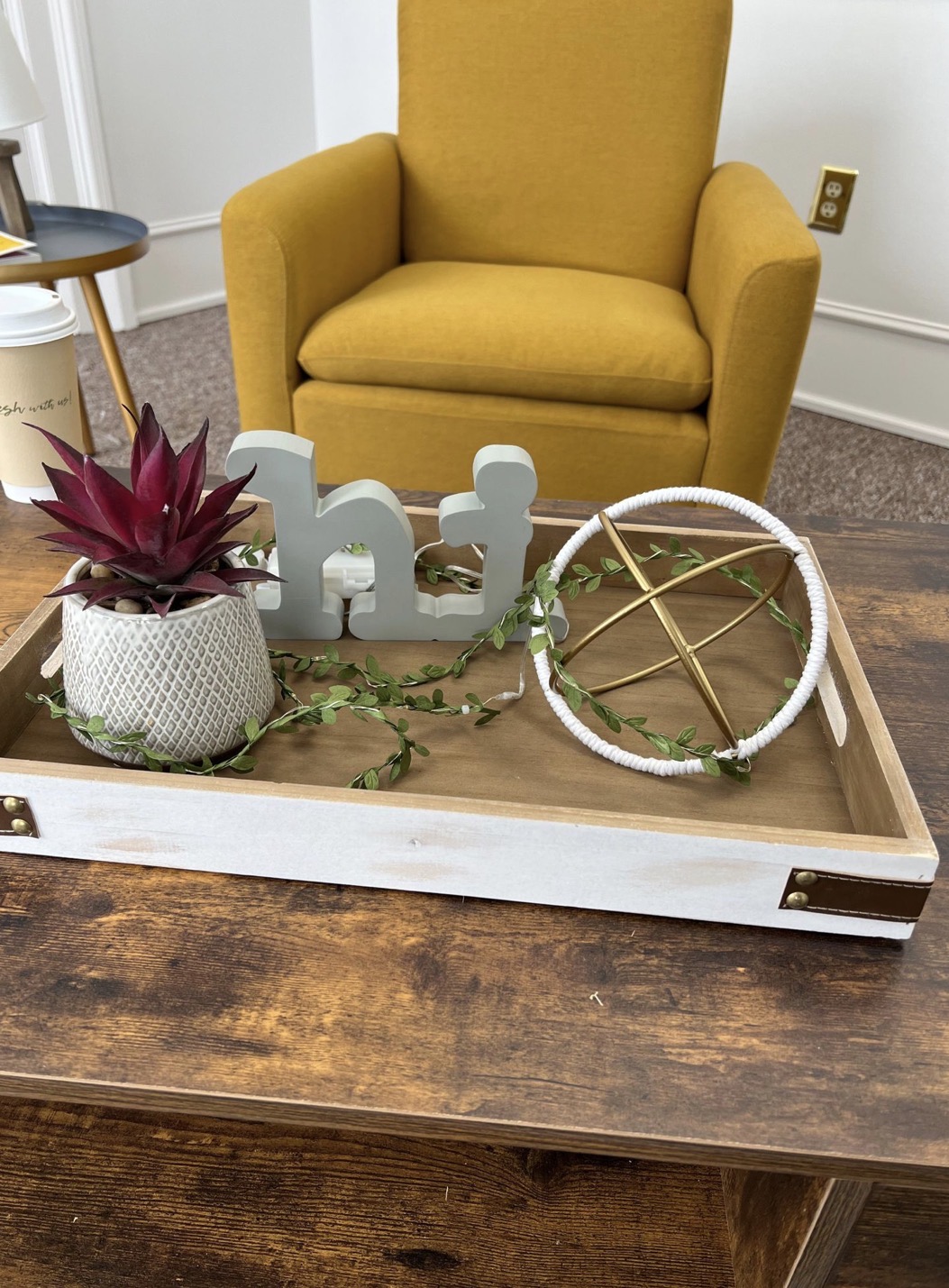 Yellow armchair behind a wooden tray with decorative items, including a potted plant, white wooden figures, and a circular wreath, on a wooden table.
