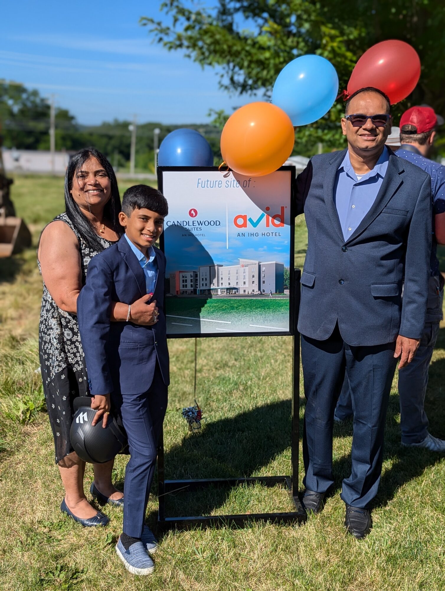 Family of three standing outdoors with balloons and a sign, smiling, on a sunny day.