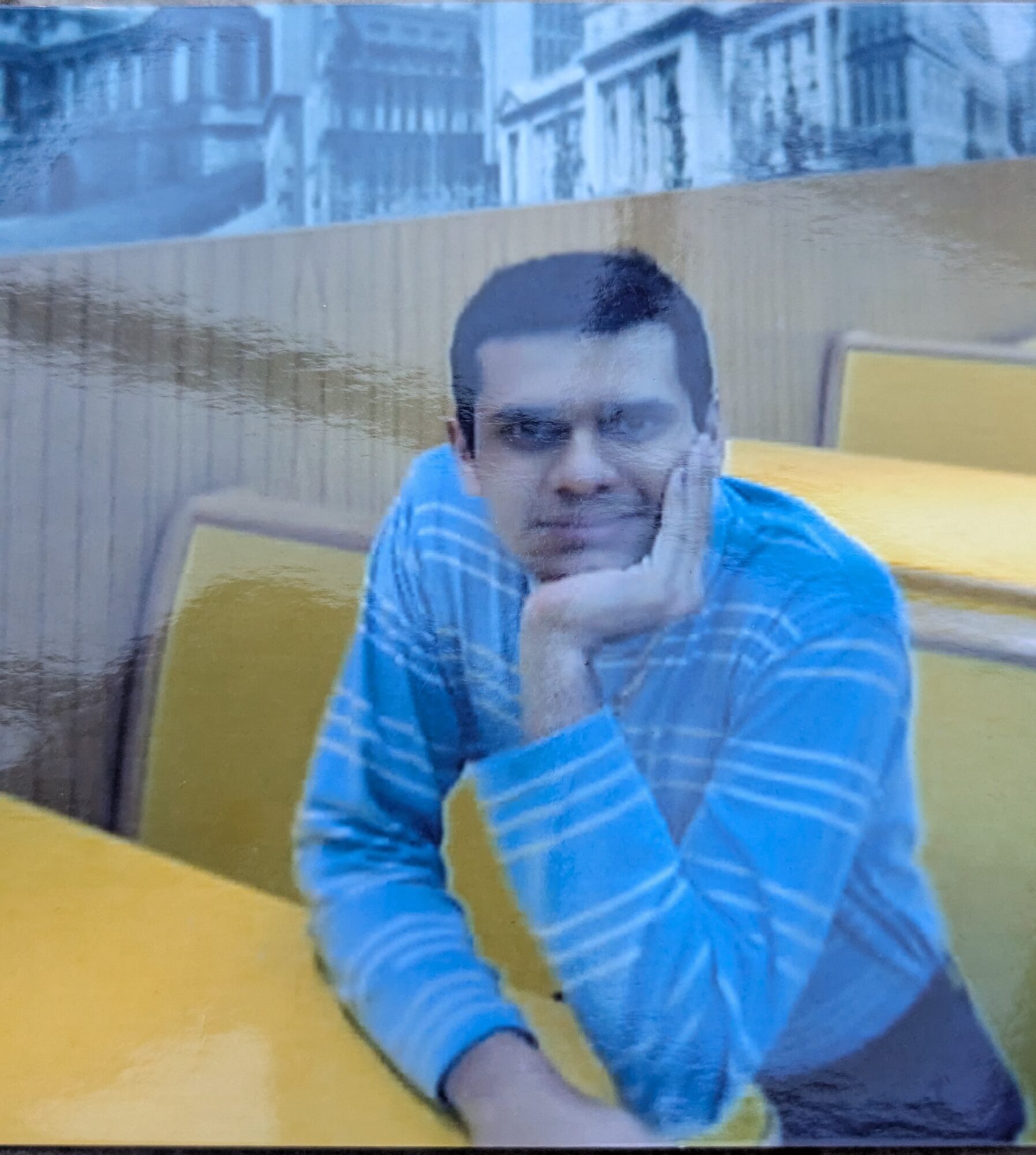Man sitting at a table with yellow chairs, resting his chin on his hand, wearing a blue striped shirt.