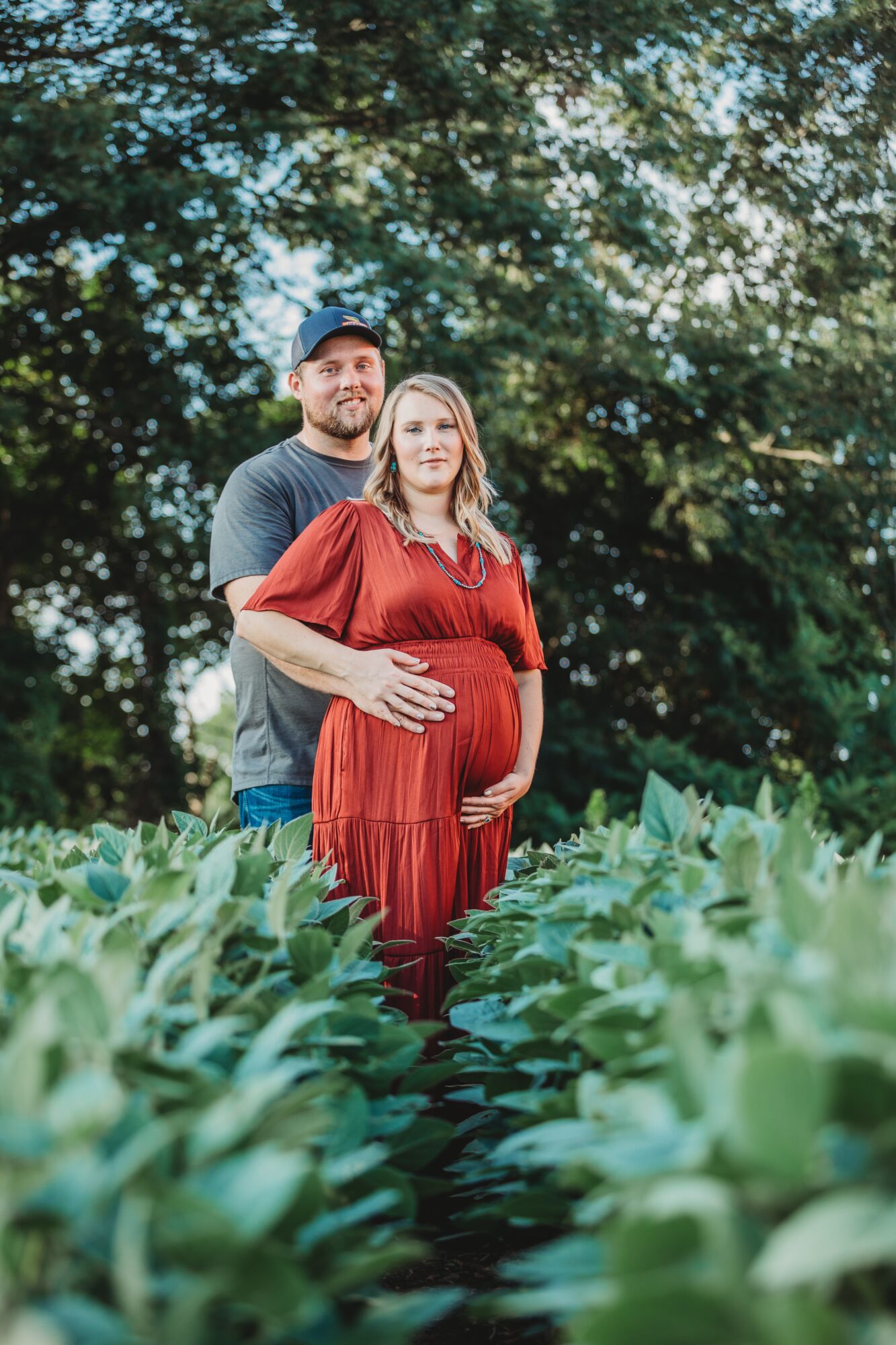 Pregnant woman in red dress standing outdoors with man behind her, surrounded by green plants and trees.