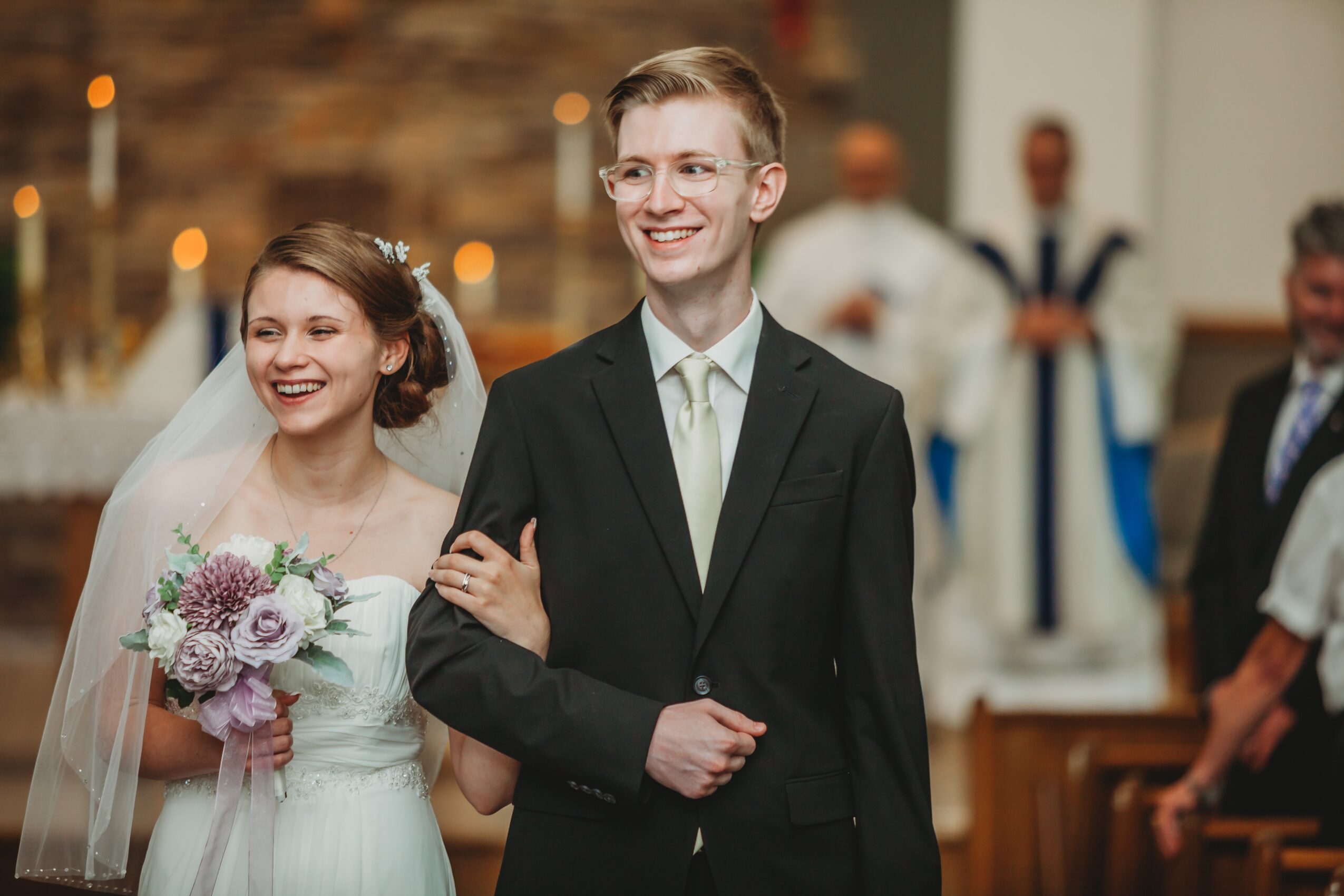 A smiling bride and groom walk arm in arm during a wedding ceremony, with guests and a priest in the background.
